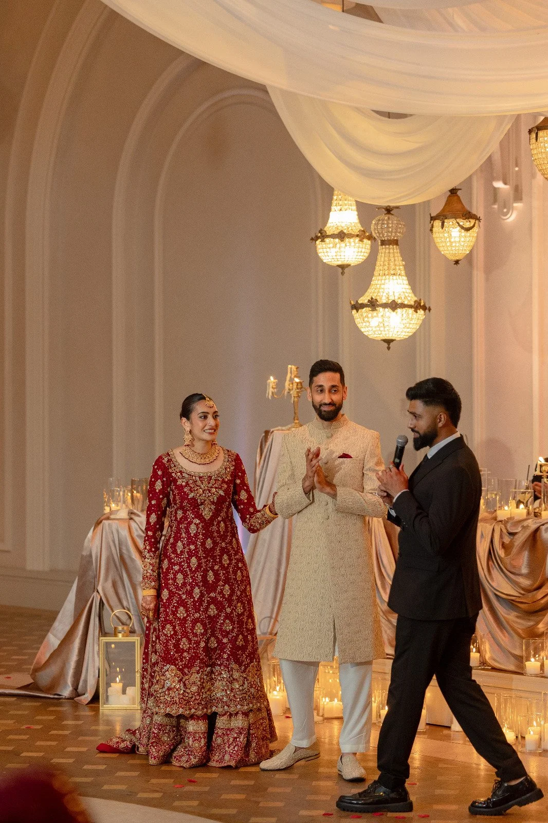 A traditional Indian wedding ceremony with a bride in a red embroidered dress, a groom in a cream sherwani, and a man in a black suit holding a microphone, all standing in a decorated hall with chandeliers, candles, and draped fabric.
