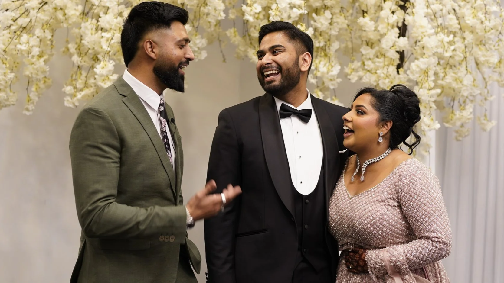 Three people at a wedding reception, two men and one woman, engaging in conversation and smiling. The man in the middle is wearing a black tuxedo, and the woman is wearing an elegant pink dress with jewelry. The background features white floral decorations.