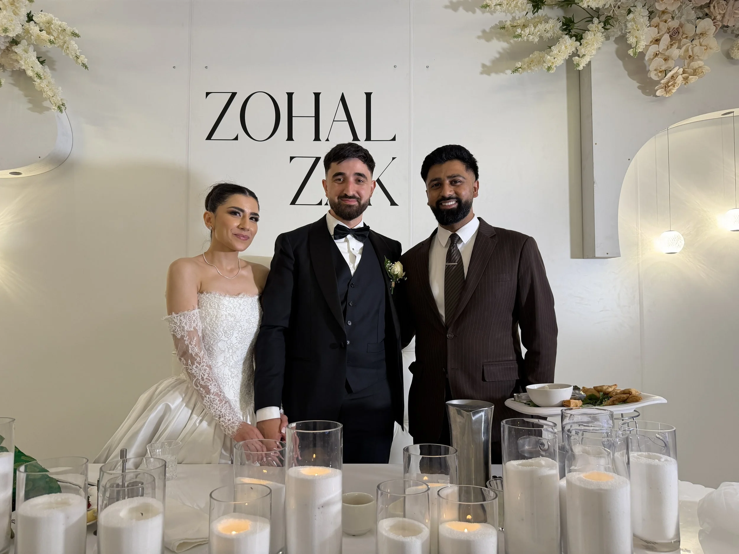 A wedding couple, with the bride wearing a wedding dress and the groom in a tuxedo, standing together with another man in a suit, at a wedding reception table decorated with lit candles and glassware.