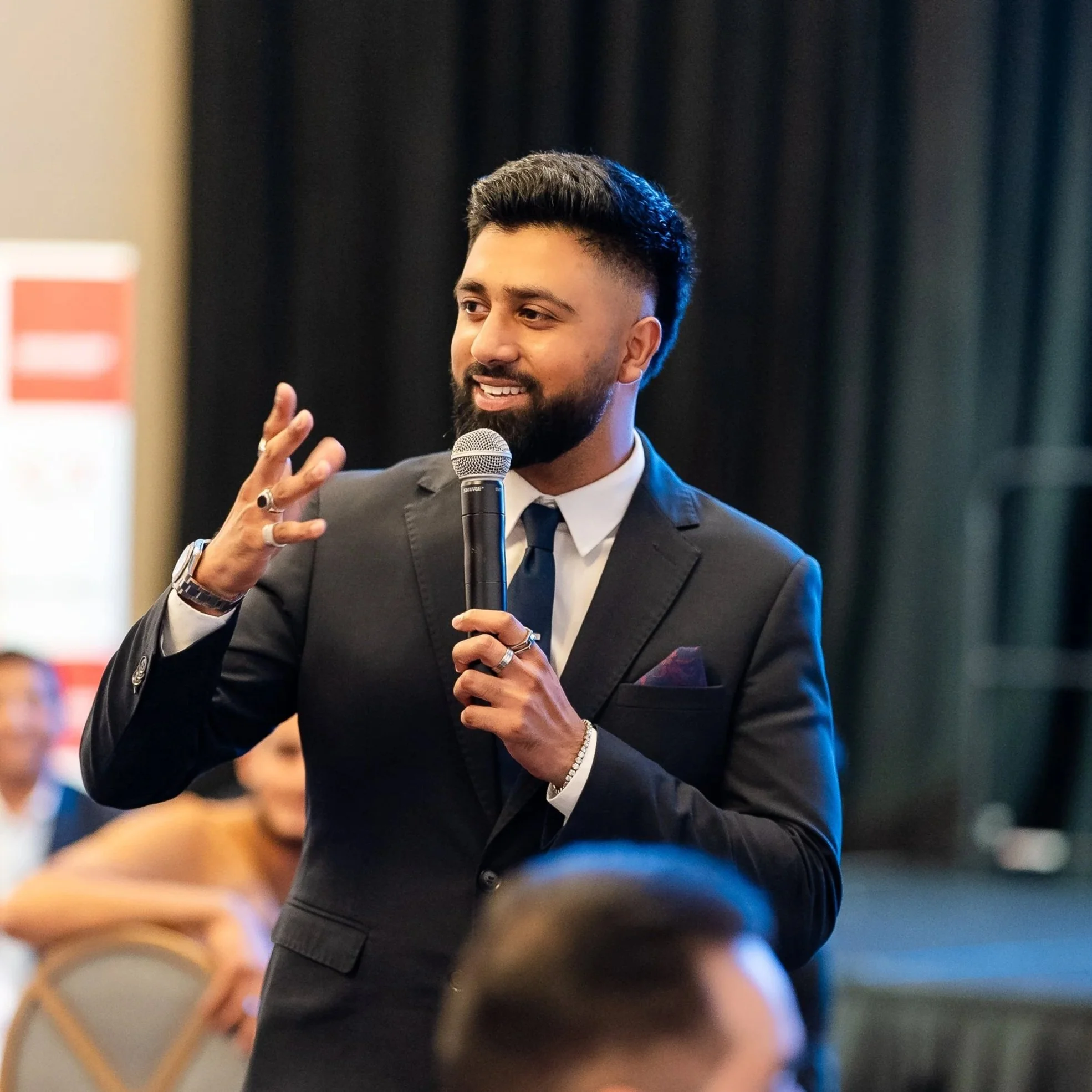 Man with dark hair and beard speaking into a microphone during a presentation, dressed in a suit and tie, in a dimly lit setting with dark curtains and blurred audience in the background.