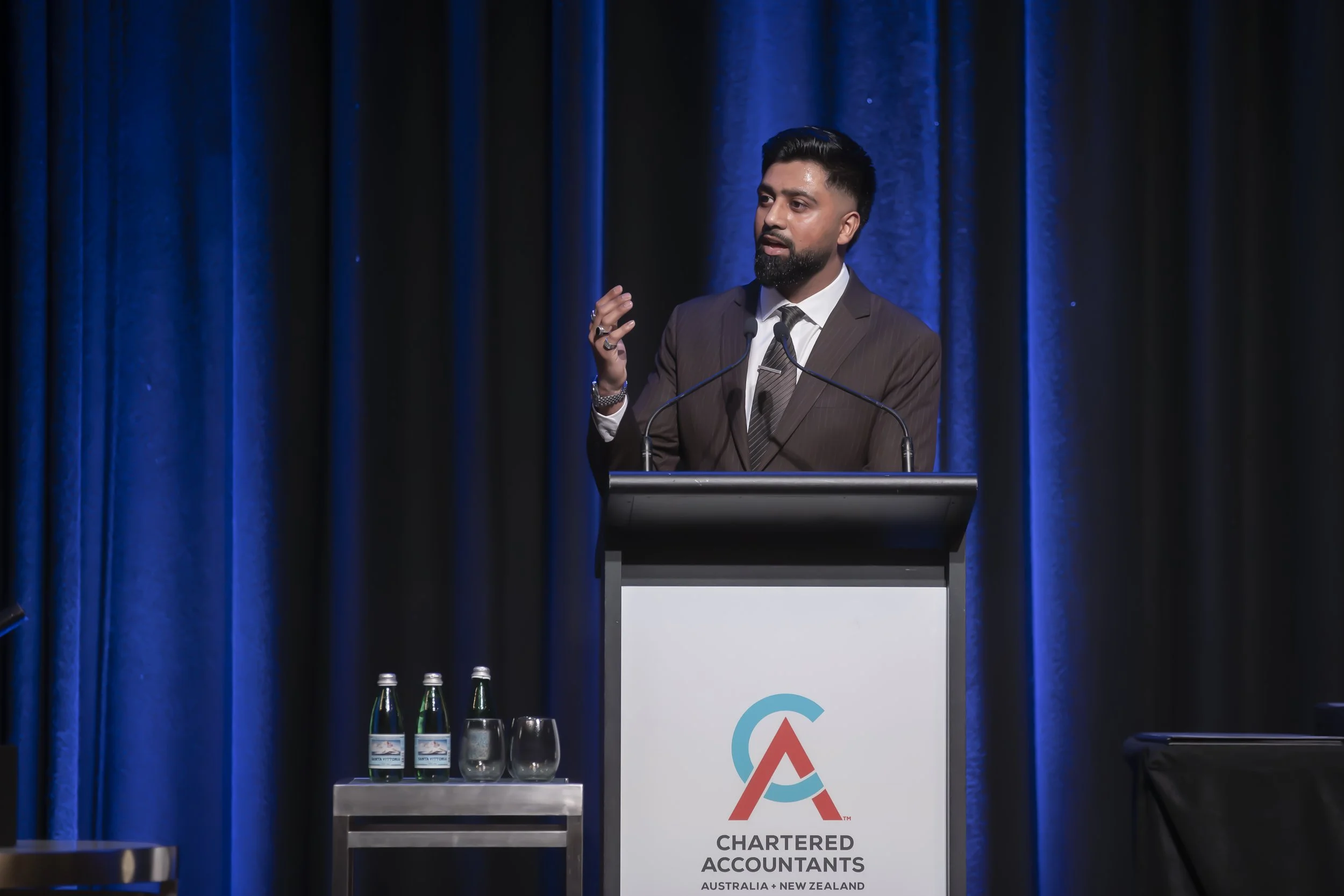 A man in a suit and tie is speaking at a podium with a microphone, in front of a dark blue curtain. The podium has a logo and the text "CHARTERED ACCOUNTANTS AUSTRALIA + NEW ZEALAND." Three bottles of water and two glasses are on a small table next to the podium.