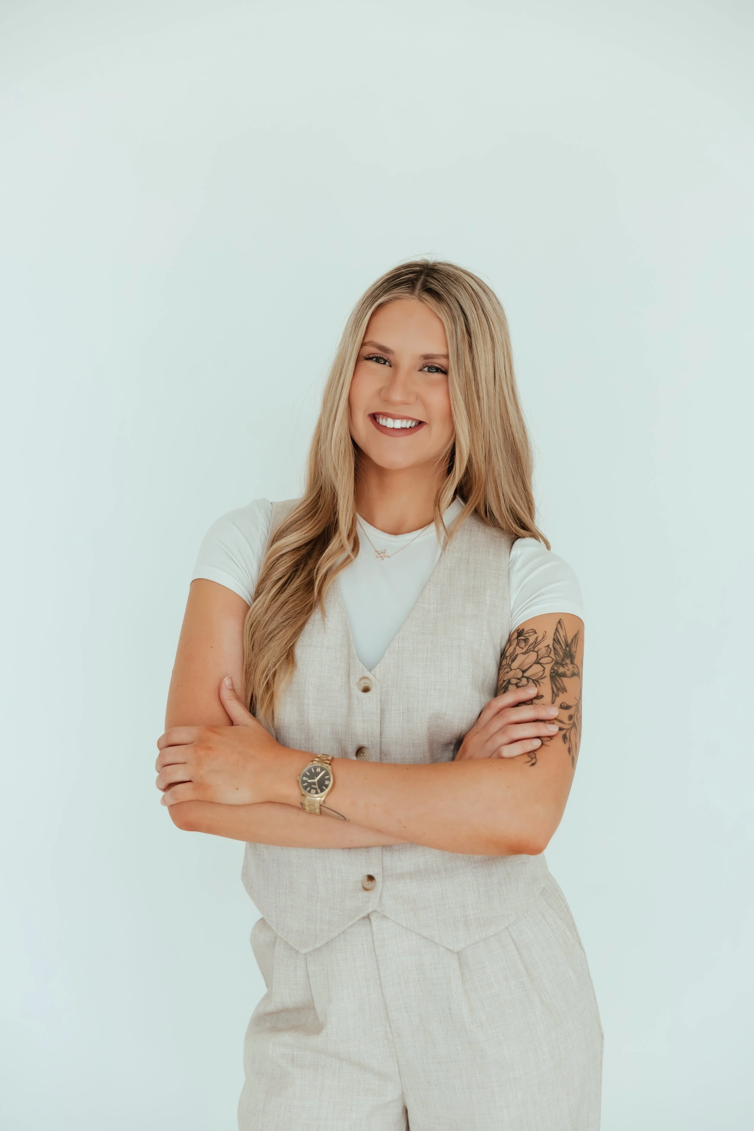 A young woman with long blonde hair smiling with her arms crossed, wearing a white t-shirt, beige vest, and matching pants, standing against a light background.