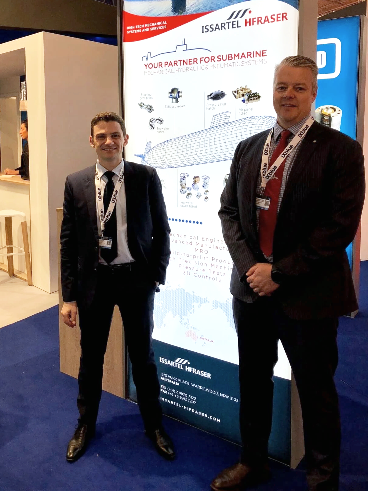 Chris Williams with a male colleague dressed in suits standing in front of a digital display at a trade show. The display promotes Issartel Hiraser, a company that provides mechanical, hydraulic, and pneumatic systems for submarines.