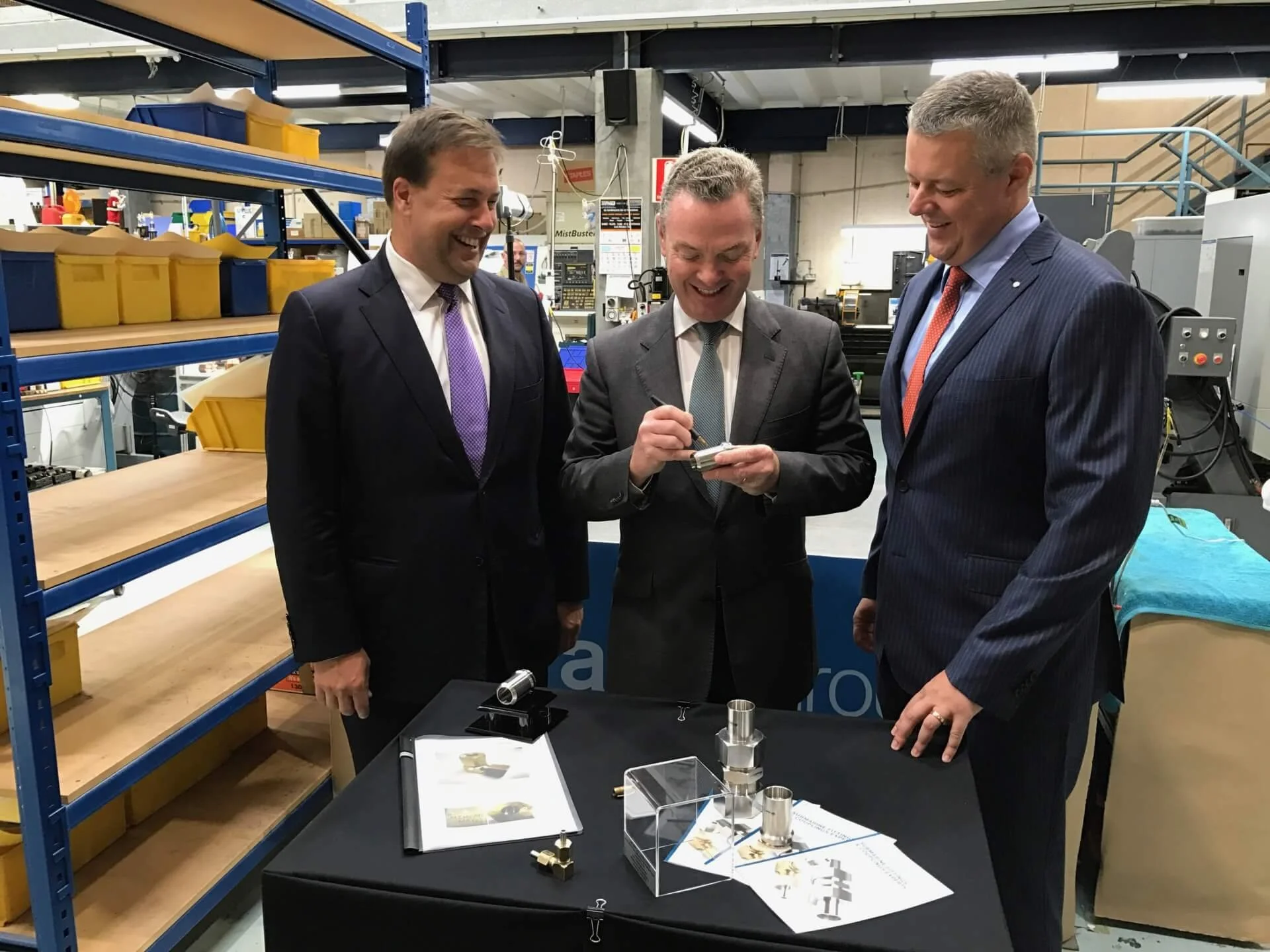 Three men in suits examining metal machine parts and technical diagrams on a table at an industrial event or manufacturing facility.