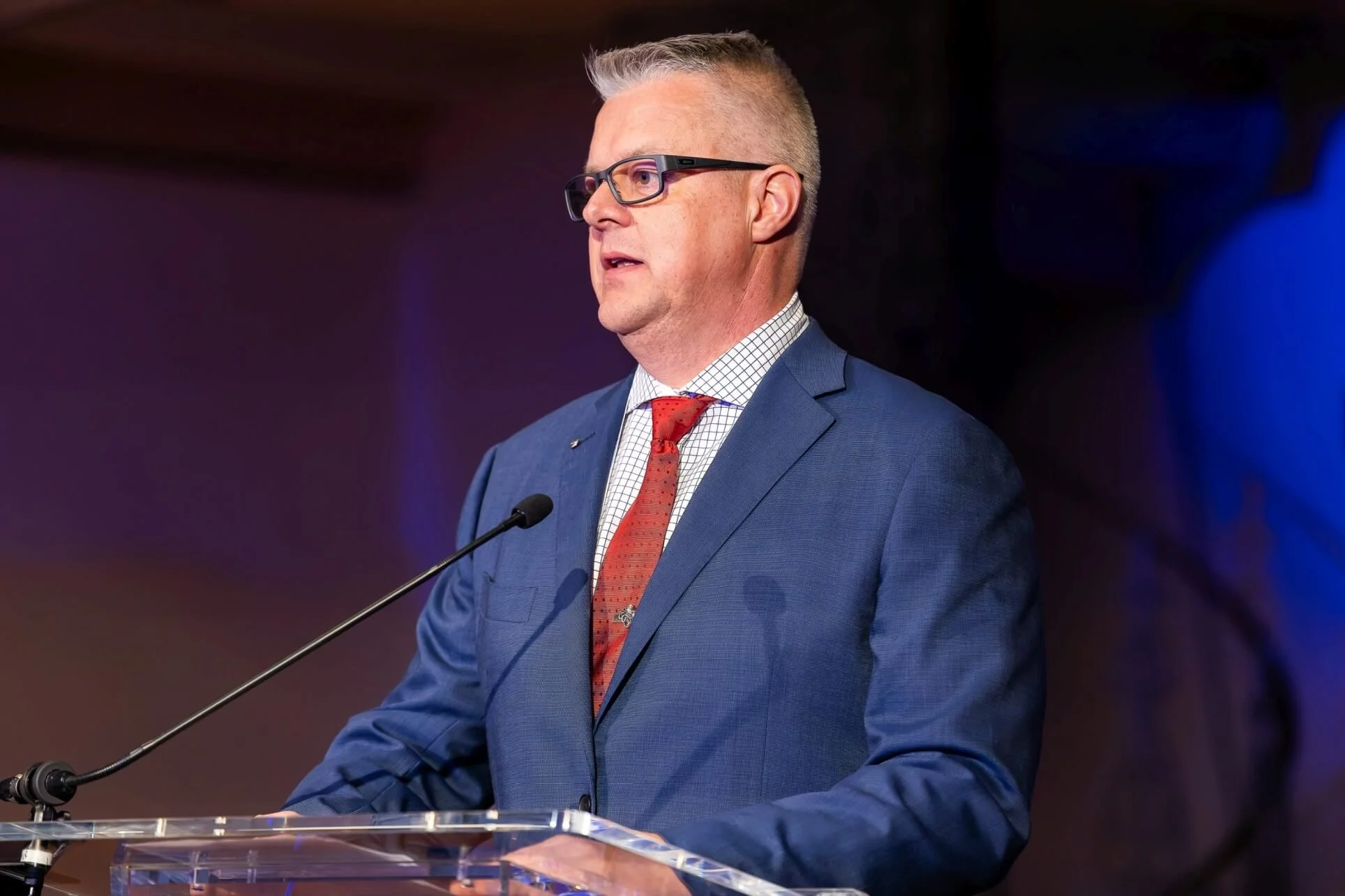 Williams Advisory with glasses stands at a podium, speaking on Australian defence industry into a microphone, wearing a blue suit, checkered shirt, and red tie, with a dark background and purple lighting.