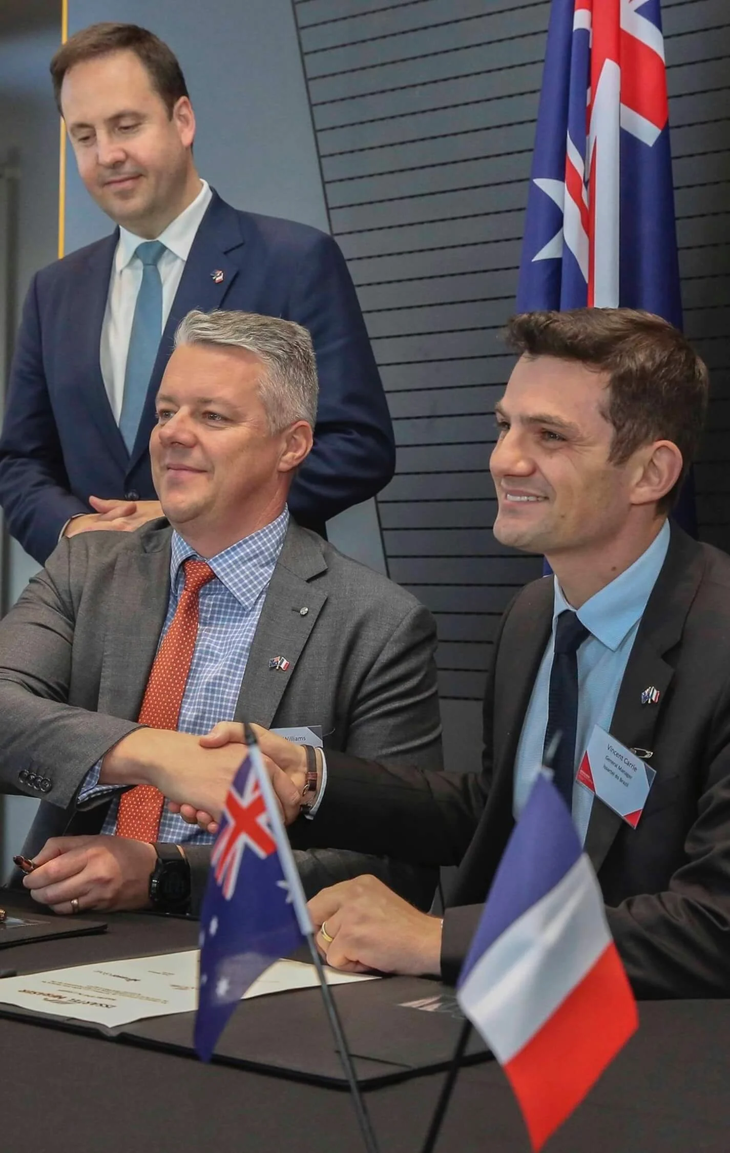Three men in business attire at a formal event, two of them shaking hands, with flags of the United Kingdom and France on the table.
