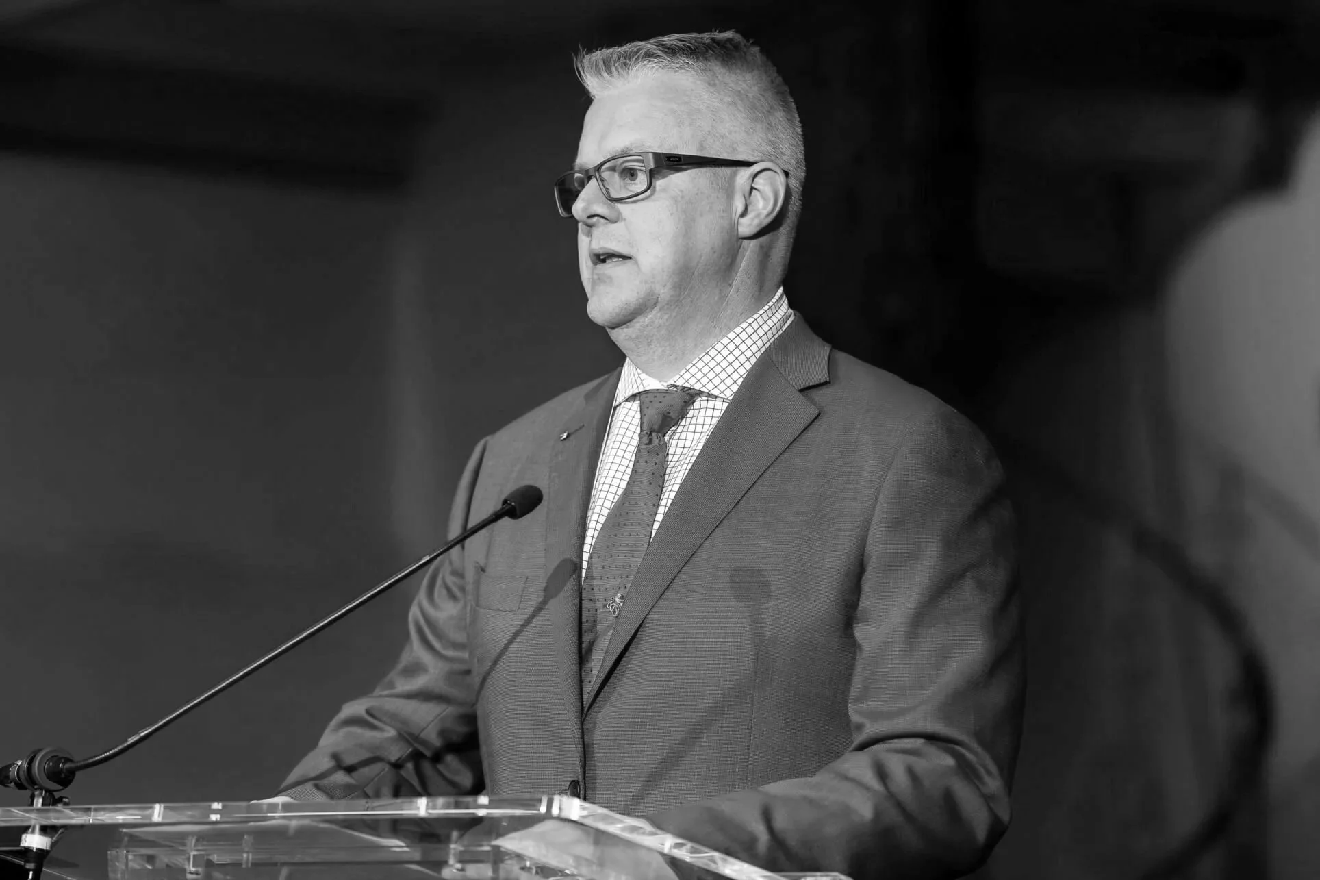 Black and white image of a Chris Williams of Williams Advisory speaking at a podium, wearing glasses and a suit with a checkered shirt and tie.
