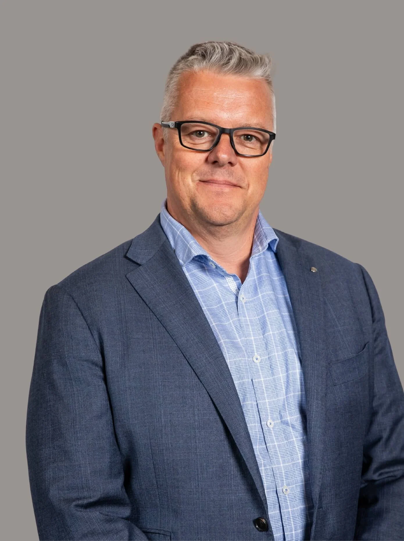 Profile colour photo of Chris Williams. Chris has short gray hair, wearing glasses, a blue blazer, and a blue checkered shirt, standing against a plain gray background.