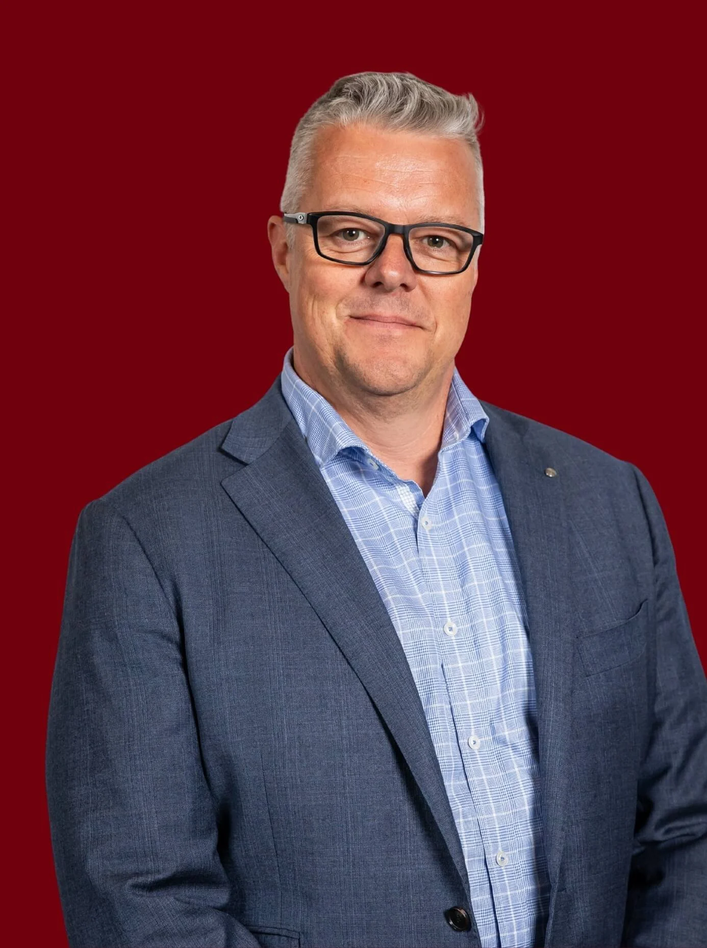 Profile colour photo of Chris Williams wearing glasses, a blue suit, and a light blue shirt, standing against a dark red background.