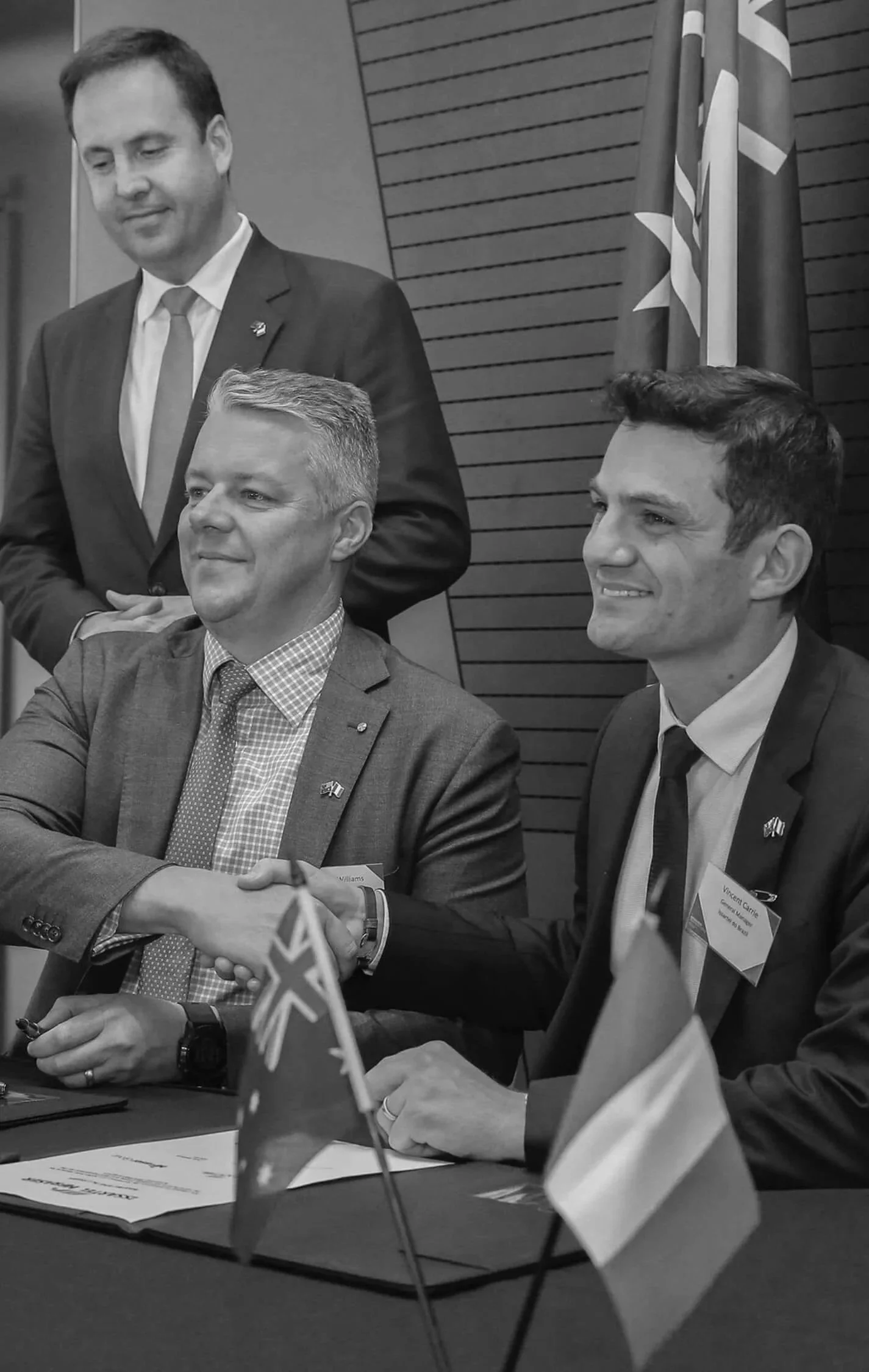 Black and white photo of Chris Williams shaking hands with Minister Ciobo shaking hands witnessing the signing of the Naval Group, with flags on the table in a professional conference room.
