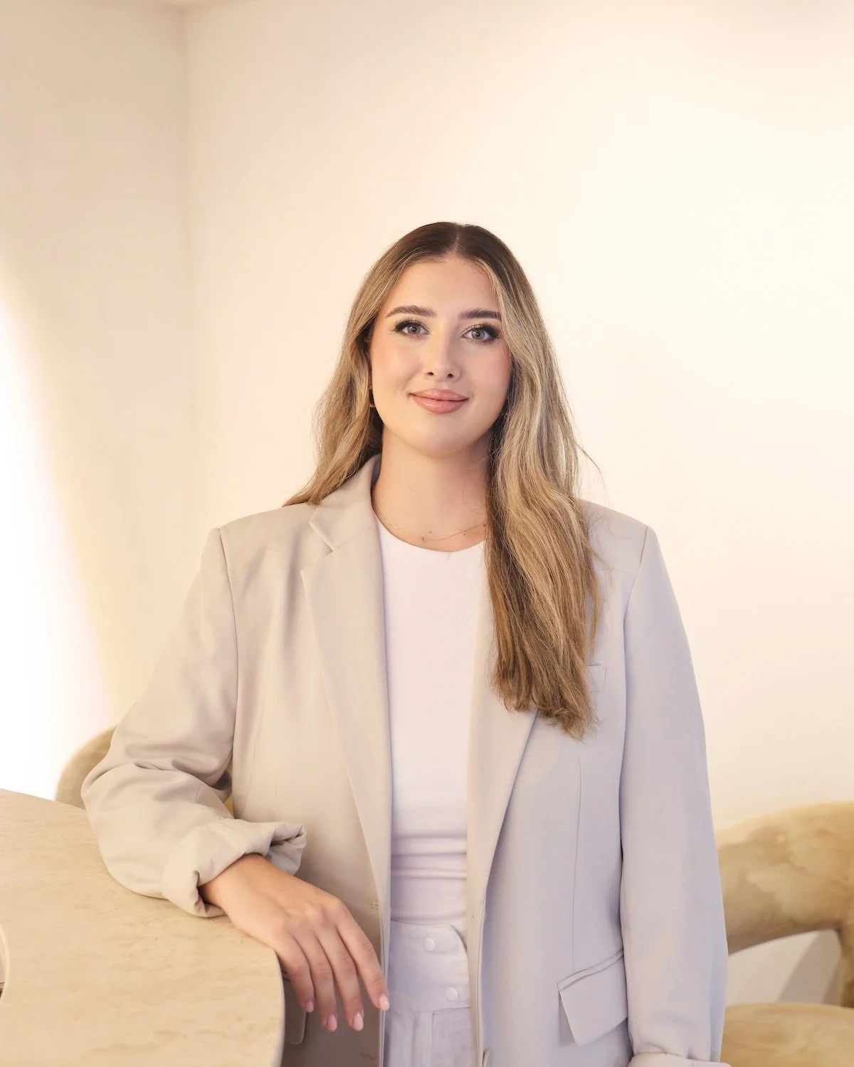A young woman with long, wavy blonde hair and blue eyes smiling, wearing a beige blazer over a white top, standing against a plain light background.