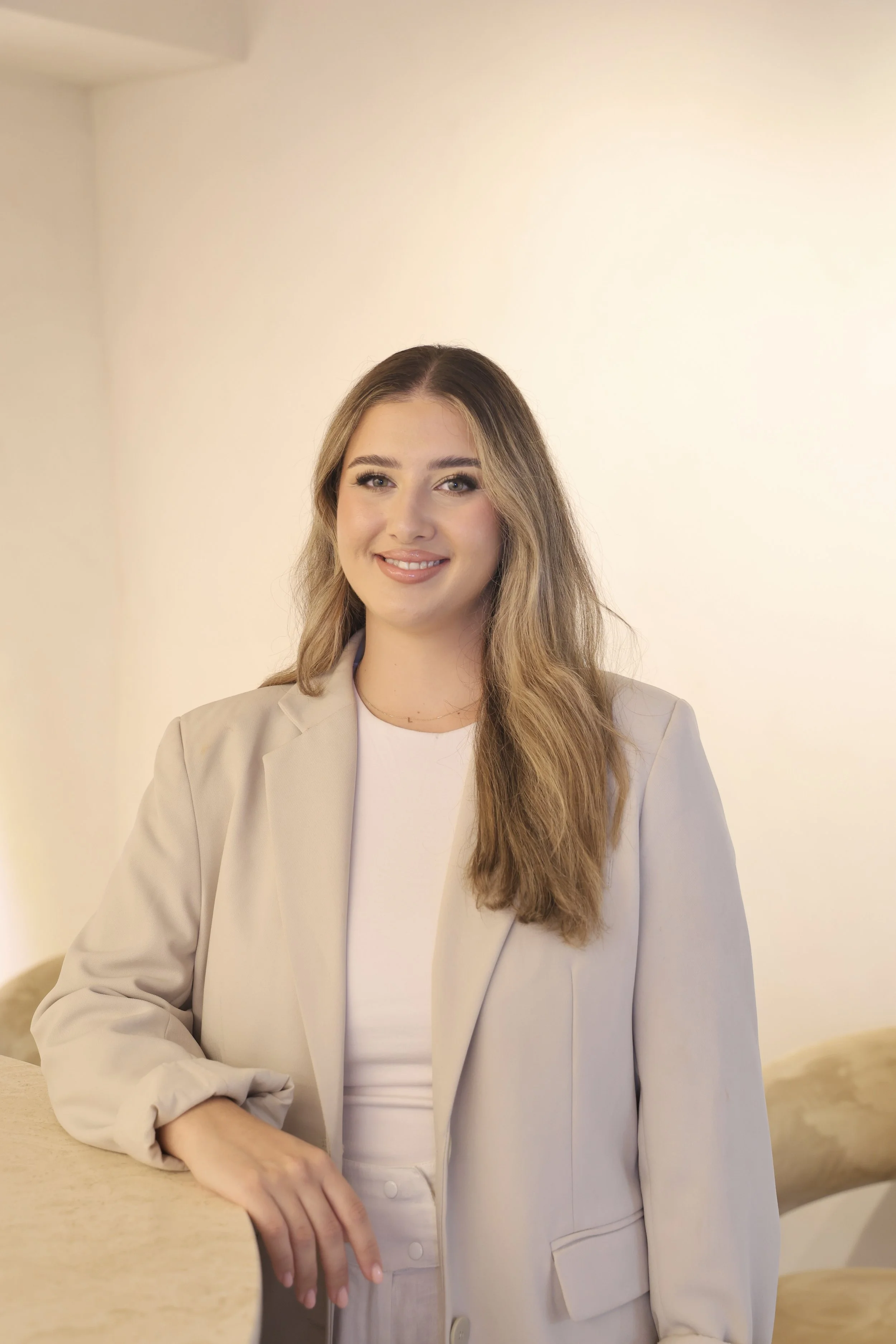 A woman with long light brown hair, wearing a beige blazer over a light top, sitting beside a wooden table with her hand resting on it, smiling, and posing indoors with a neutral background.