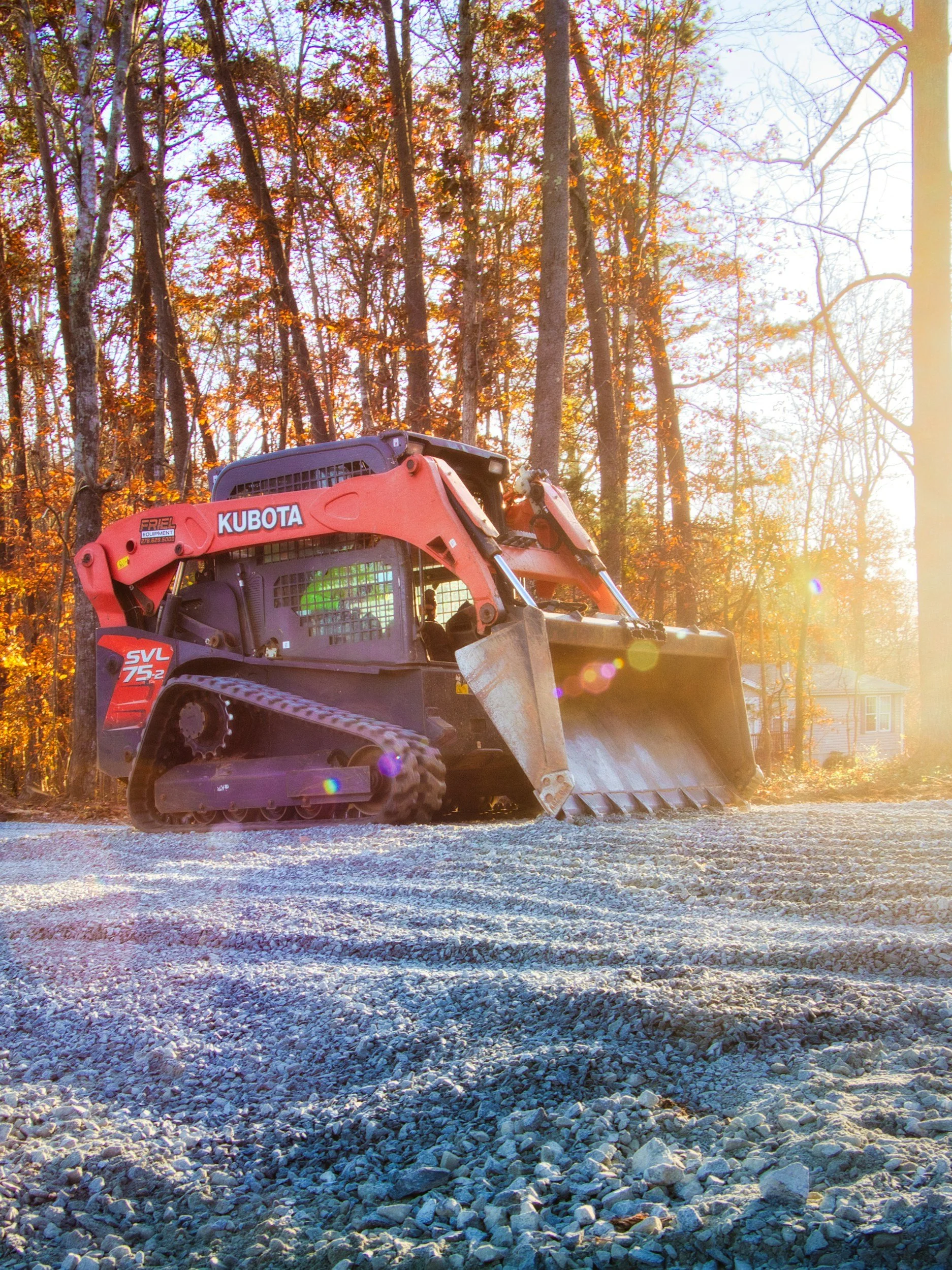 A small orange excavator with a bucket attached is working on a gravel surface with a forest of trees with orange leaves in the background, illuminated by sunlight.