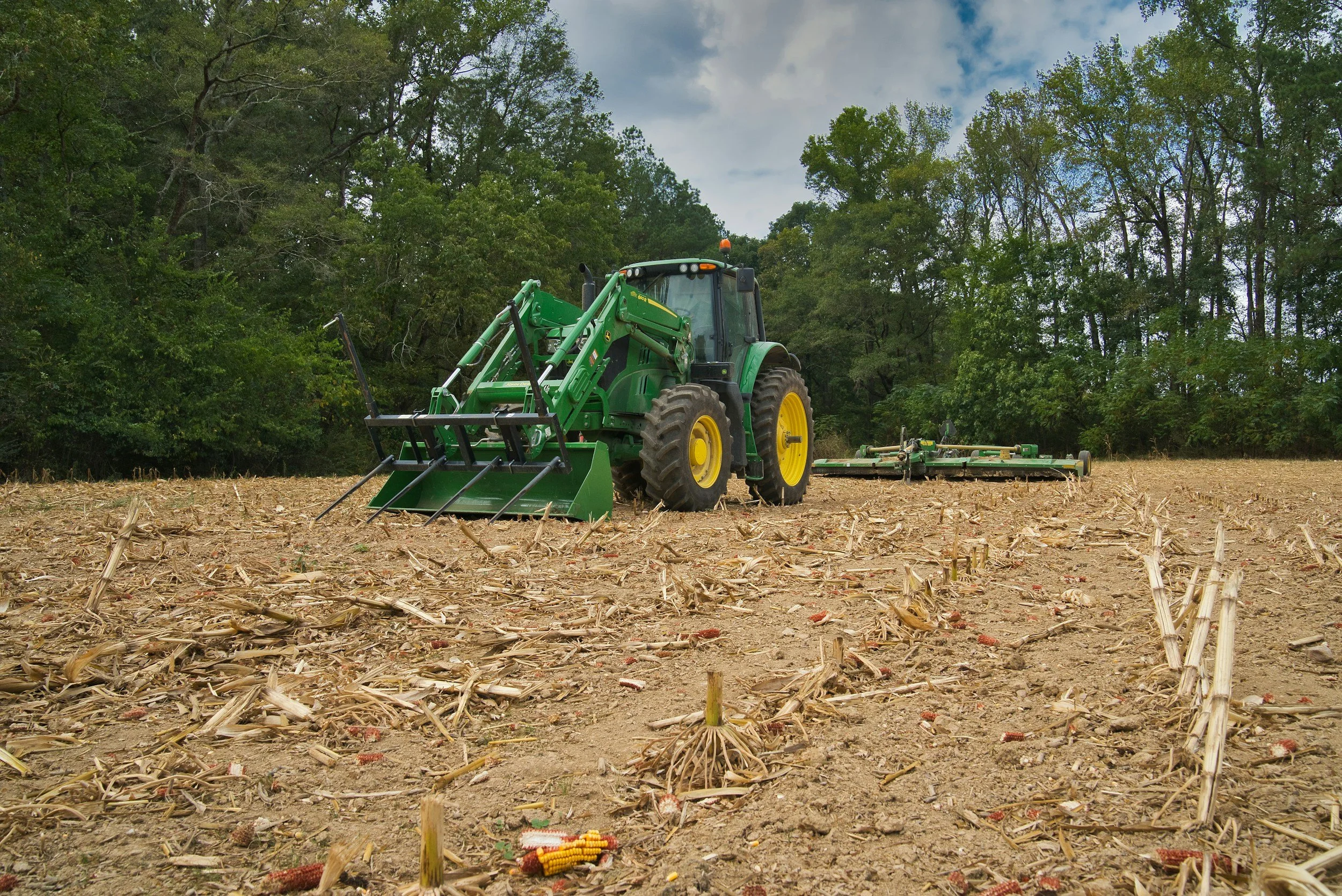 A green tractor with a front loader attachment working in a field with corn stubble, surrounded by trees under a partly cloudy sky.