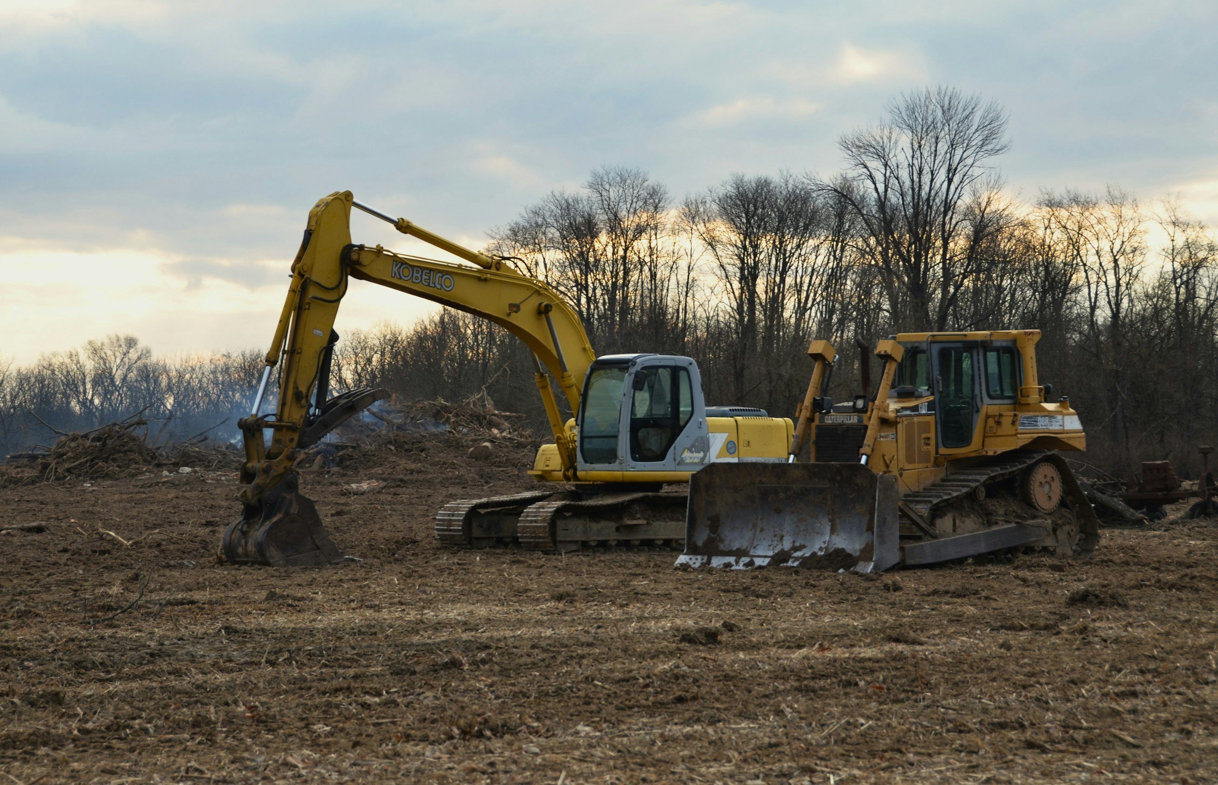 Construction site with a yellow excavator and a bulldozer working on dirt soil, with leafless trees and a cloudy sky in the background.