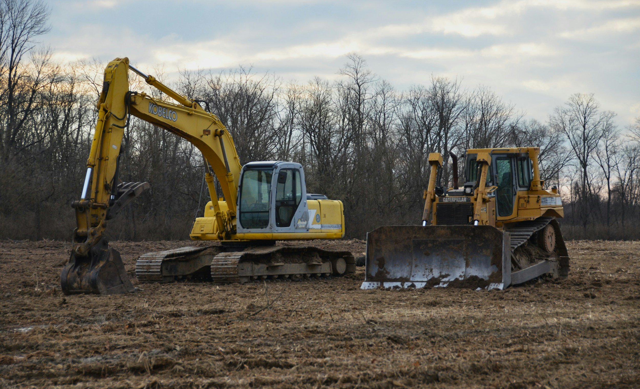 Construction site with a yellow excavator and a yellow bulldozer on dirt ground, with leafless trees in the background under a cloudy sky.