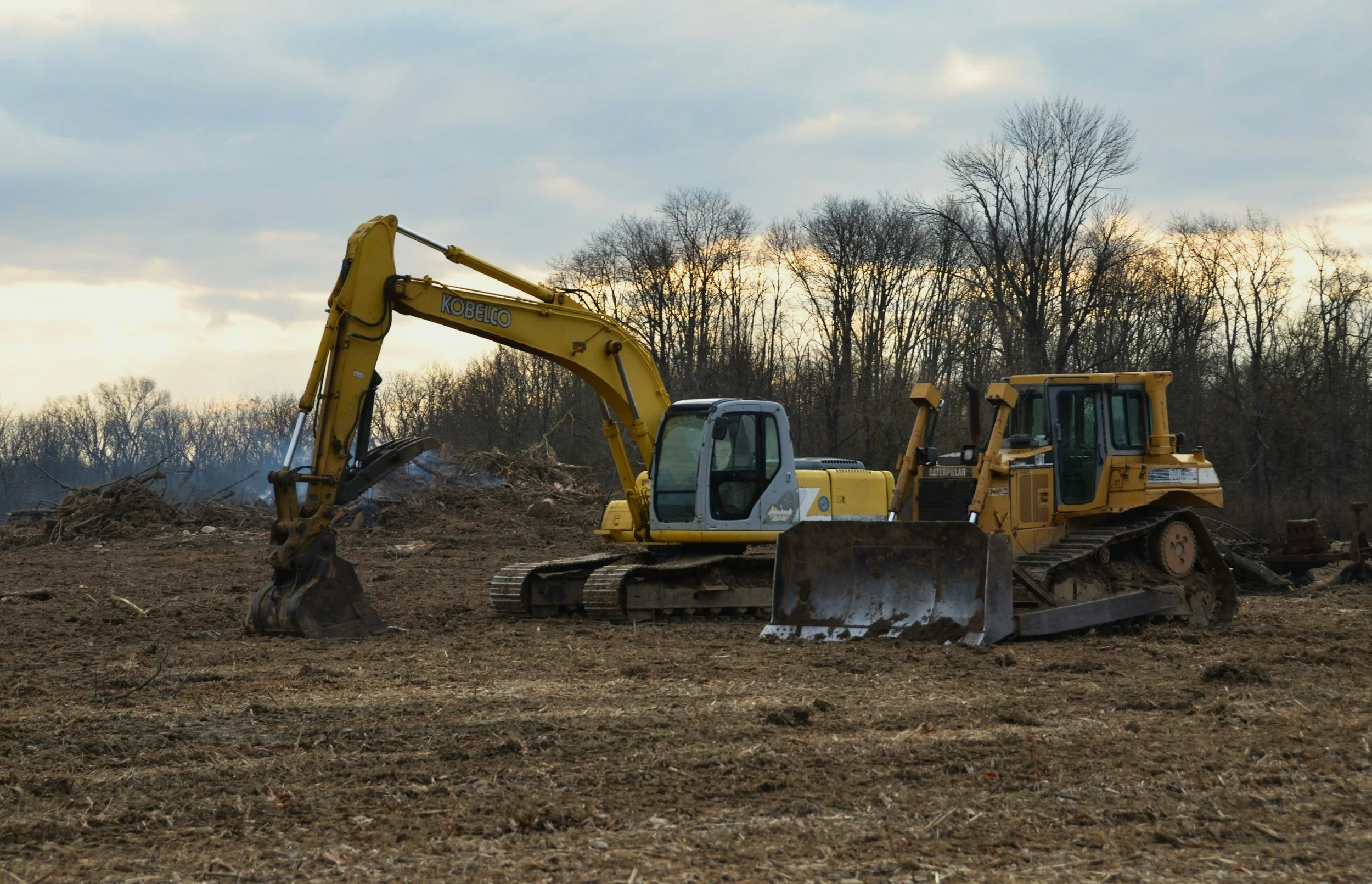 A construction site with two yellow excavators working on the dirt, with a background of leafless trees and a cloudy sky during sunset.