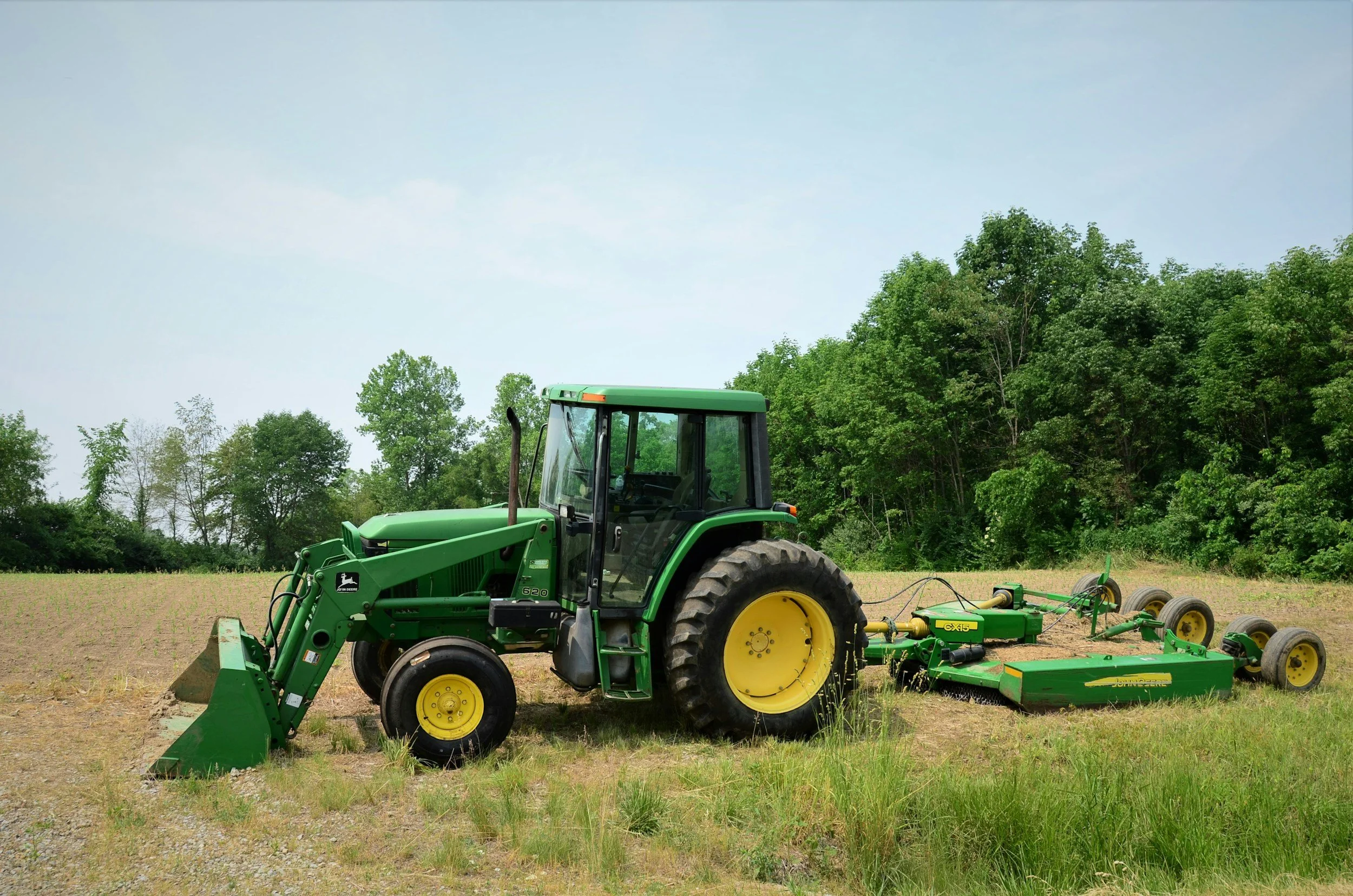 Green John Deere tractor with attached farming equipment on an open field, trees in the background, under a partly cloudy sky.