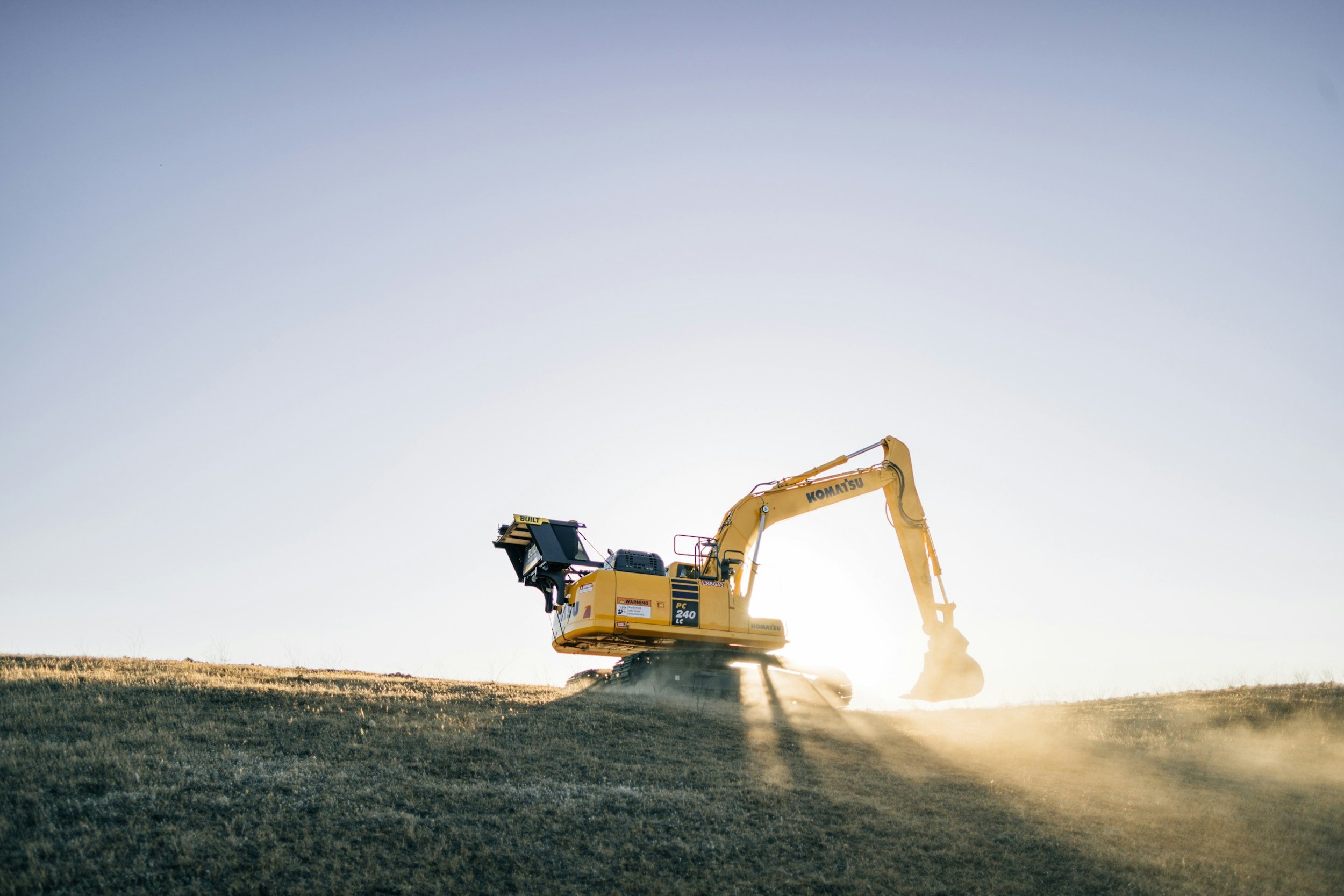 A yellow Komatsu excavator operating on a grassy field during sunset, with dust rising from the ground.