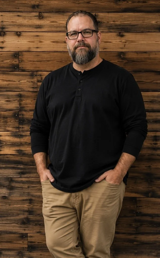 A man with glasses and a beard wearing a black long-sleeve shirt and khaki pants standing with hands in pockets in front of a wooden wall.