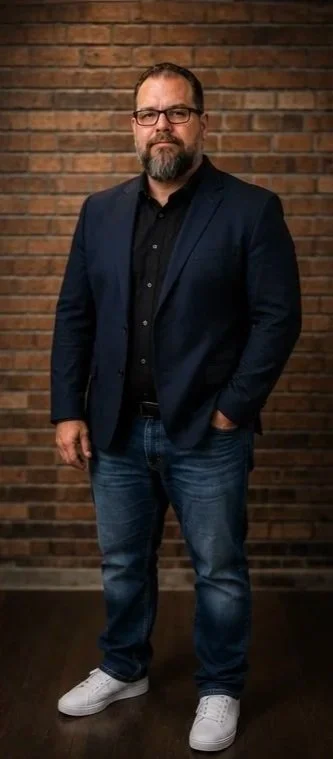 A man with glasses and a beard wearing a dark blazer, black shirt, jeans, and white sneakers standing in front of a brick wall.