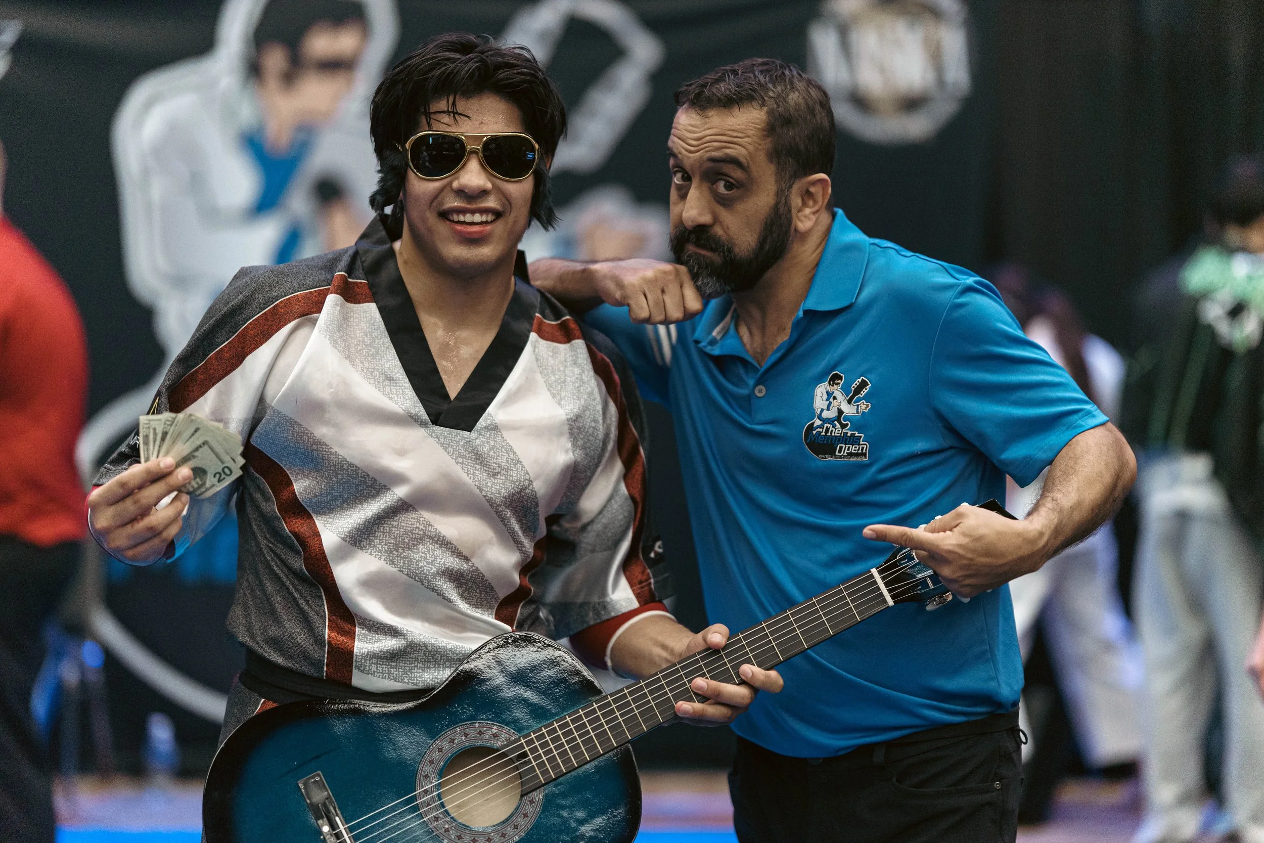 Two men stand close together, one holding a guitar and a stack of cash, with someone playing in the background. The man on the left wears sunglasses and a patterned sports jersey, smiling at the camera. The man on the right has a short beard, looks serious, and wears a blue polo shirt with a logo for the Memphis Open.