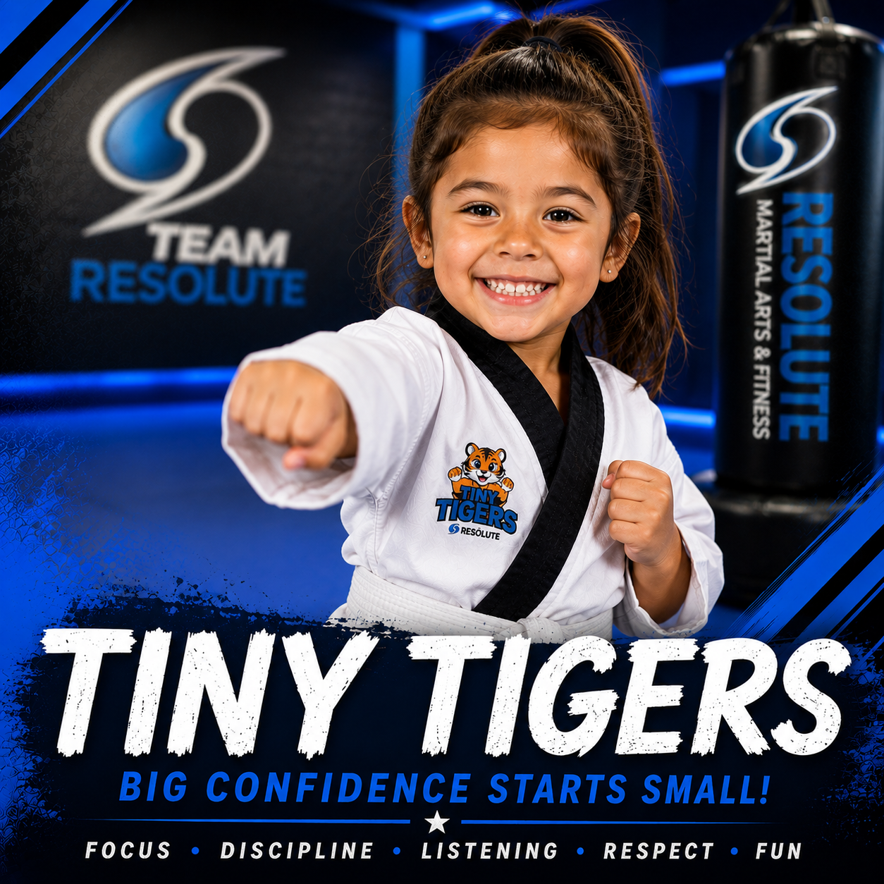 A young girl in a white martial arts uniform with a Tiny Tigers logo, smiling and posing with a fist extended in a martial arts studio with Team Resolute branding in the background.