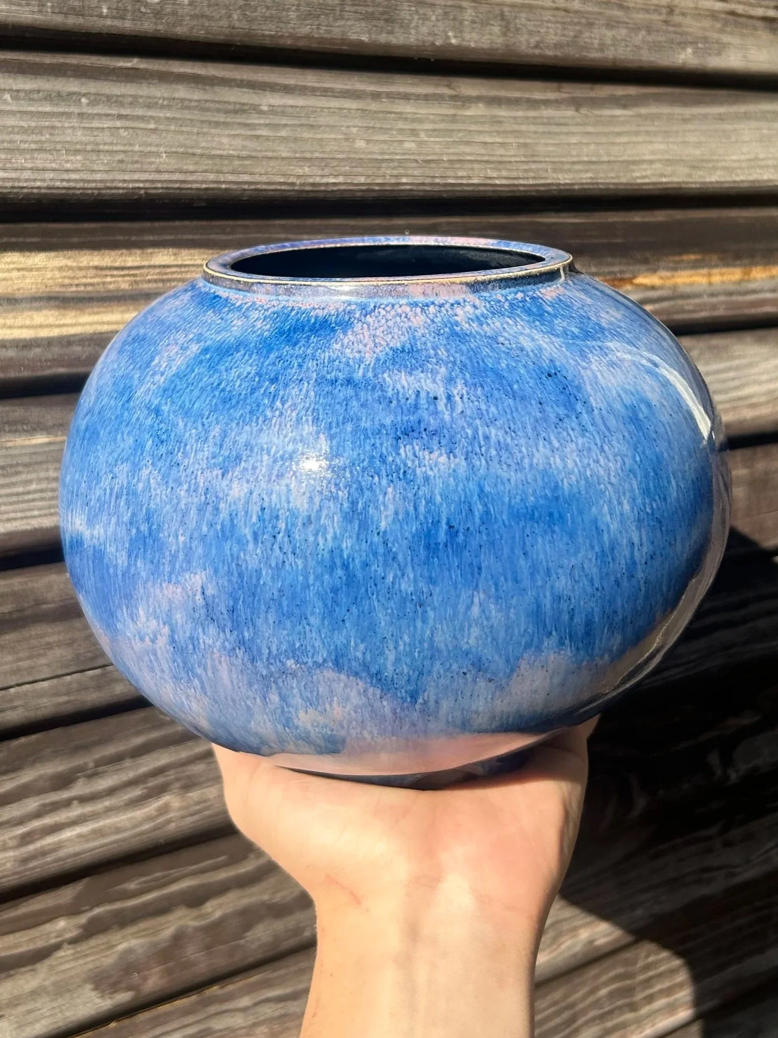 A person holding a round blue ceramic vase in front of a wooden background.