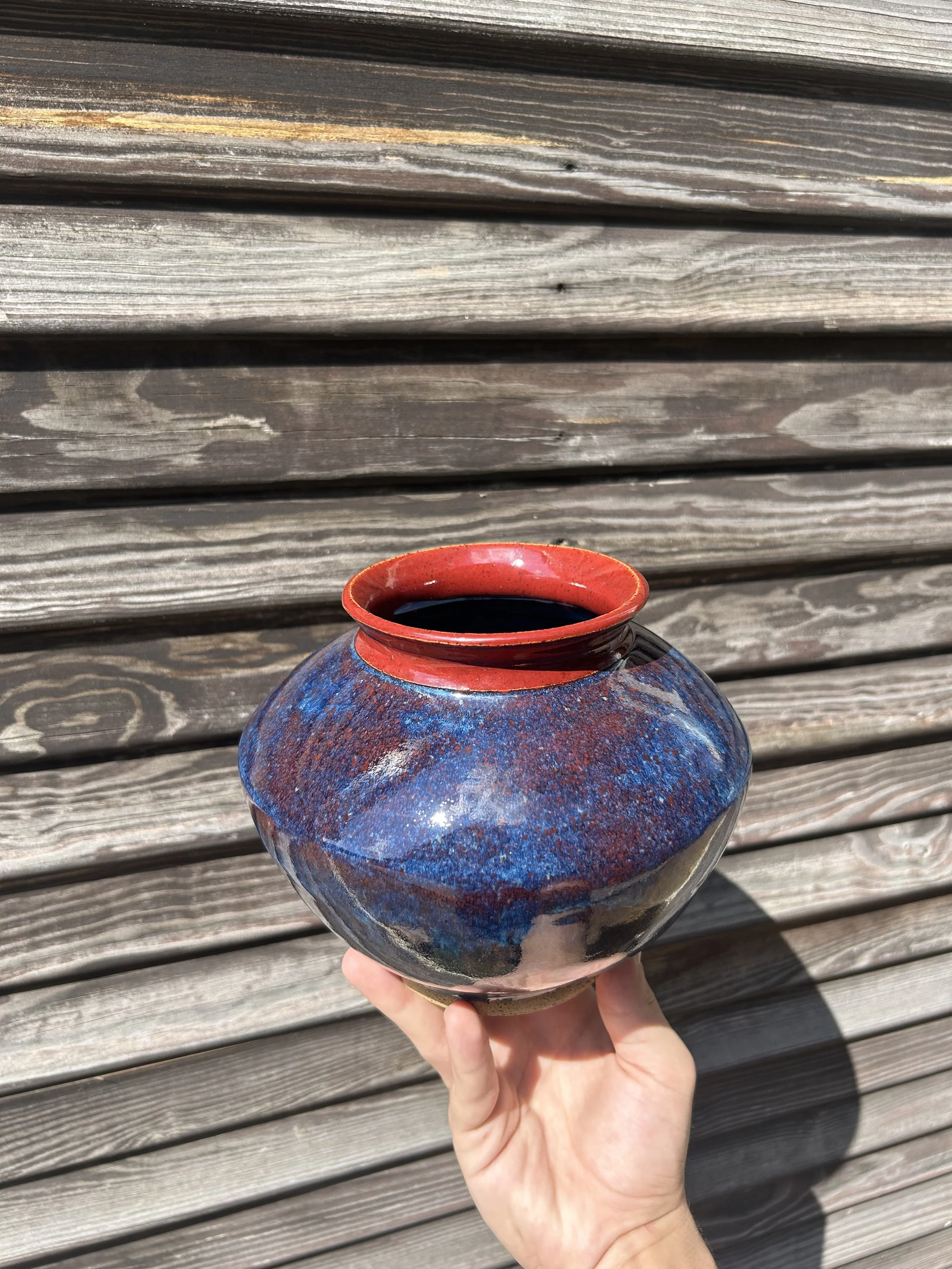 Hand holding a colorful ceramic vase with a narrow red rim against a wooden background.