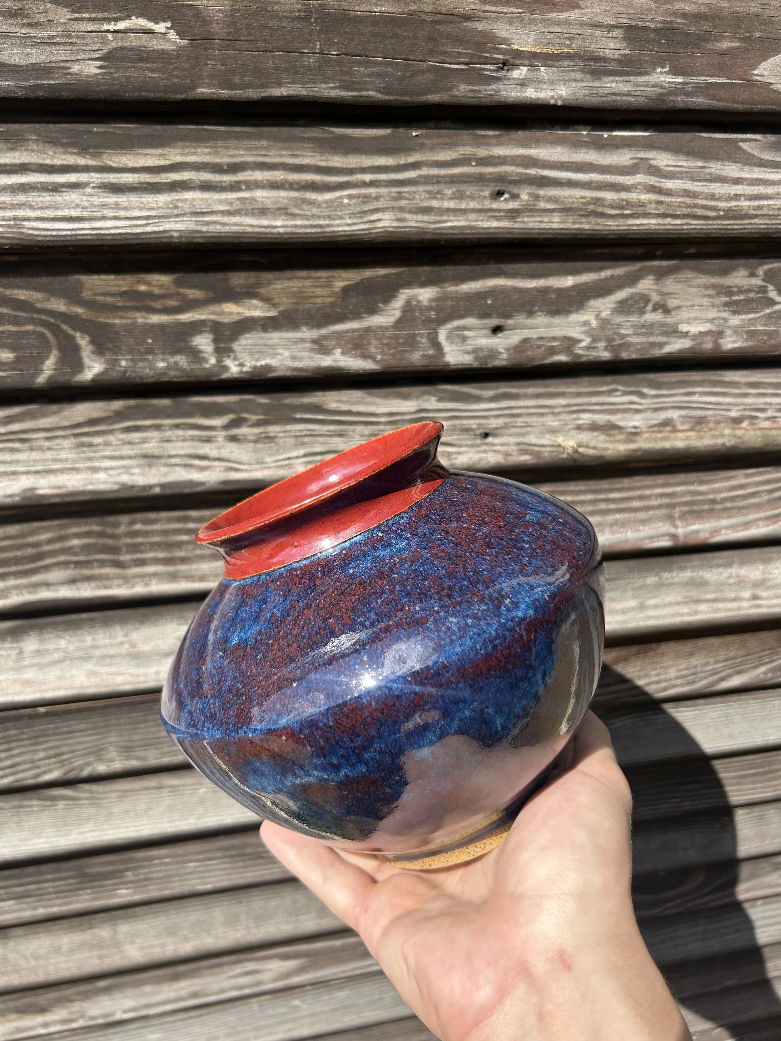A hand holding a small rounded glass sculpture with a swirling pattern of red and blue colors, set against a wooden background.