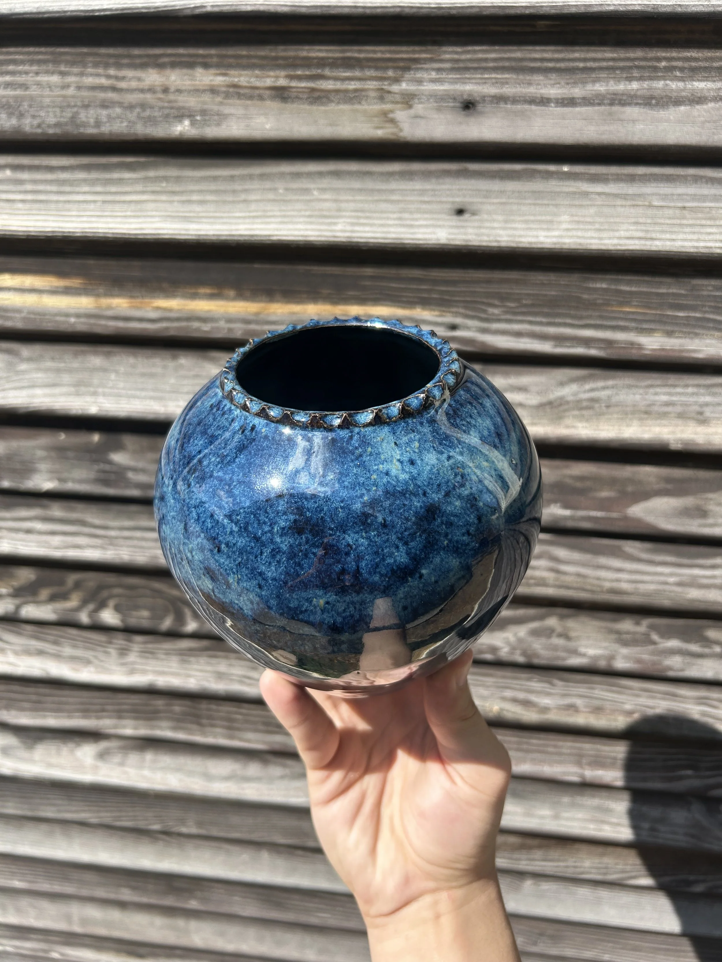 A person's hand holding a blue and gray ceramic vase with a round body and a narrow opening, set against a weathered wooden background.