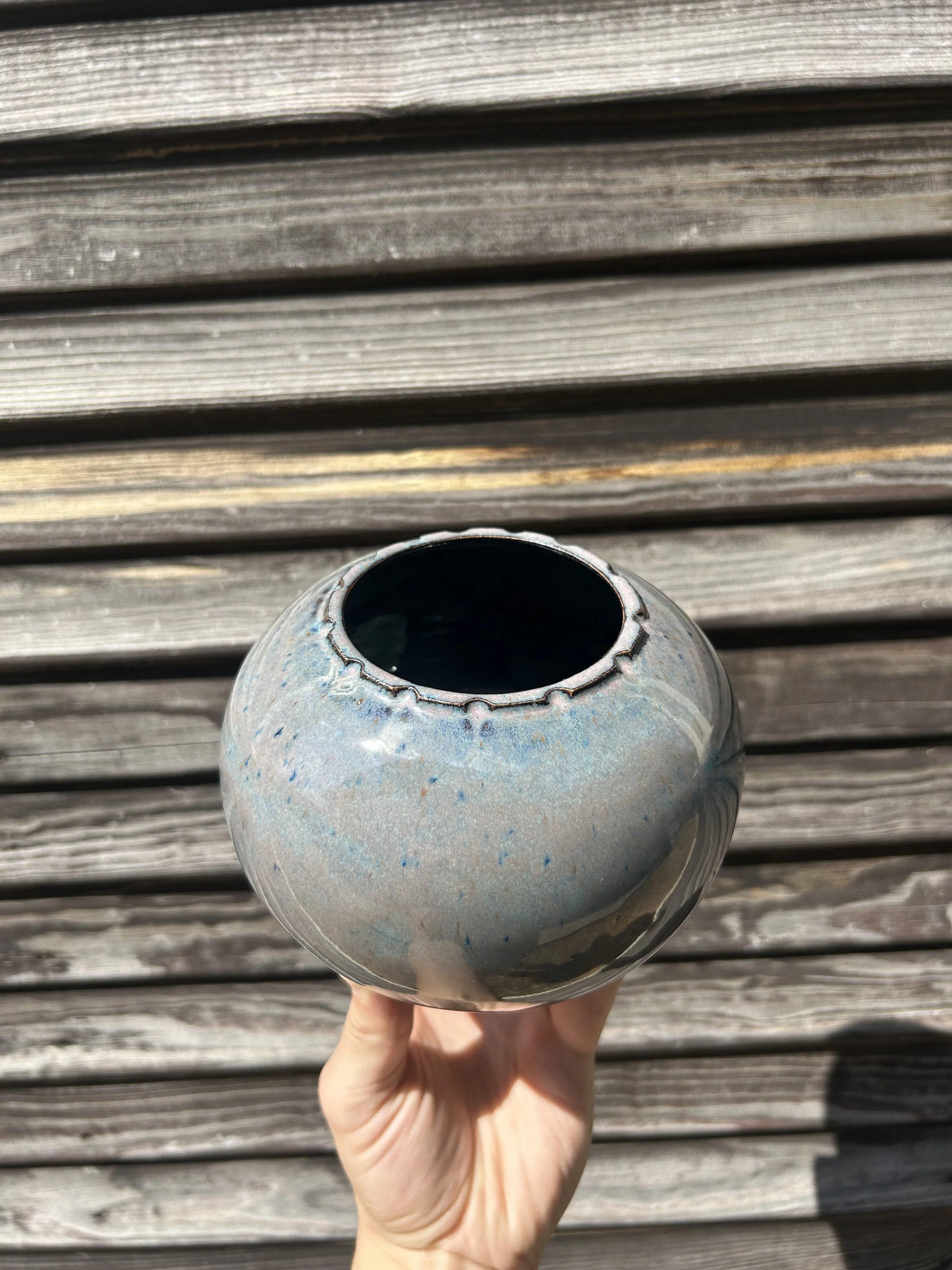 A hand holding a ceramic vase with a narrow opening, against a weathered wooden surface.