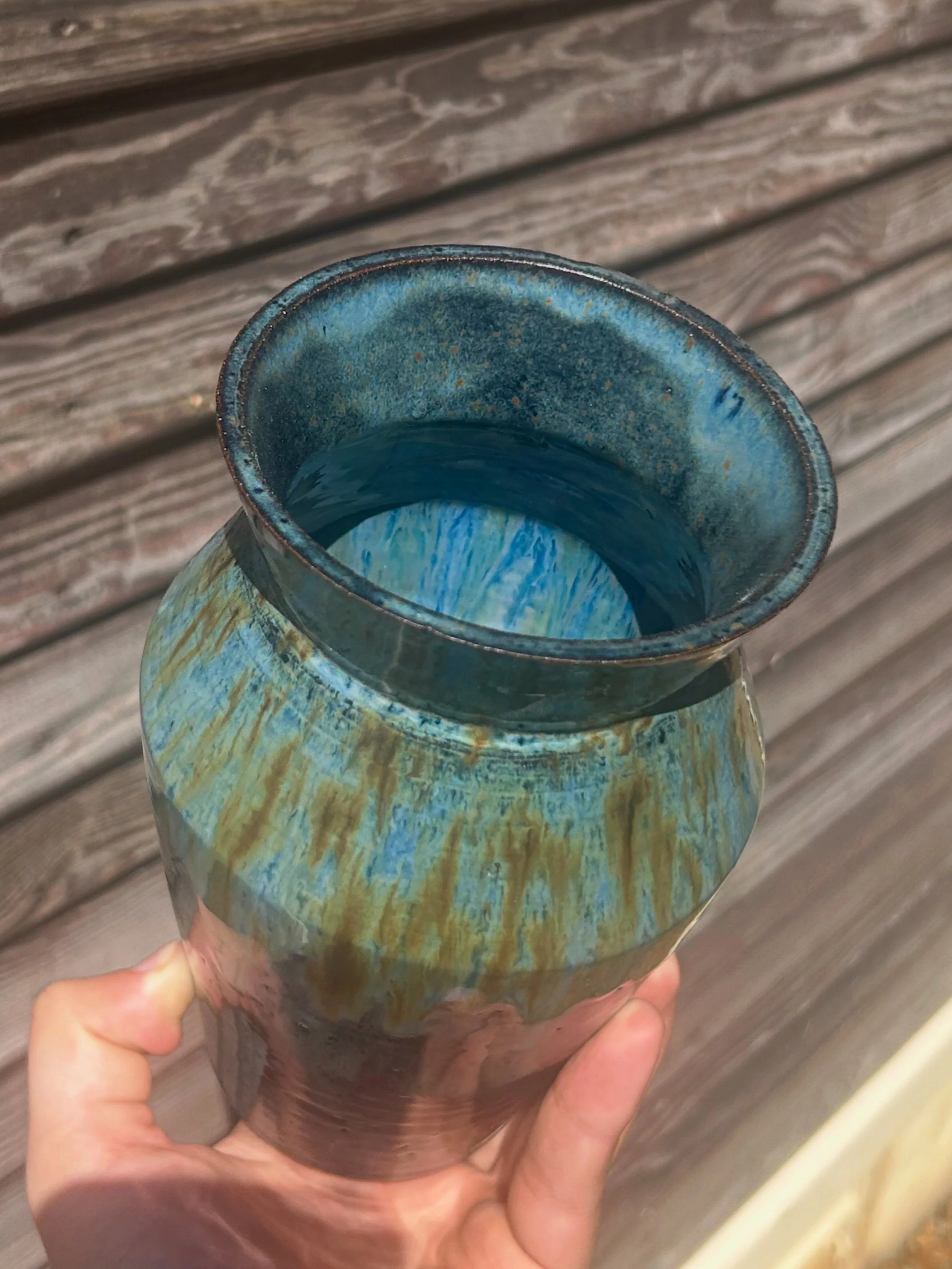 Close-up of a someone holding a ceramic vase with a rough, textured outside in shades of blue and tan, and a smooth, glossy interior in a darker blue, against a weathered wooden background.