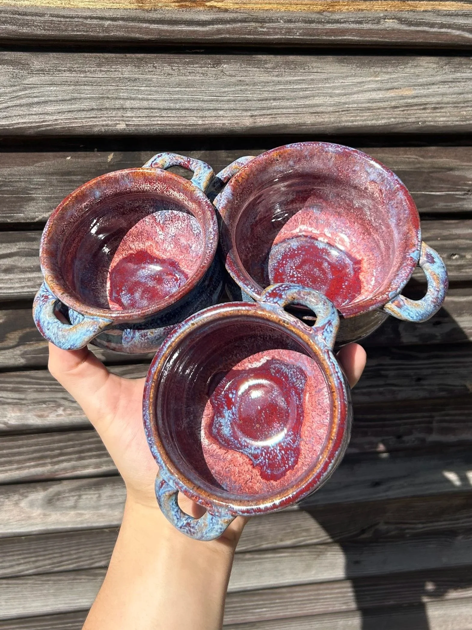 Four small ceramic cups with handles, featuring a reddish, brown, and blue glaze, held over a wooden surface.