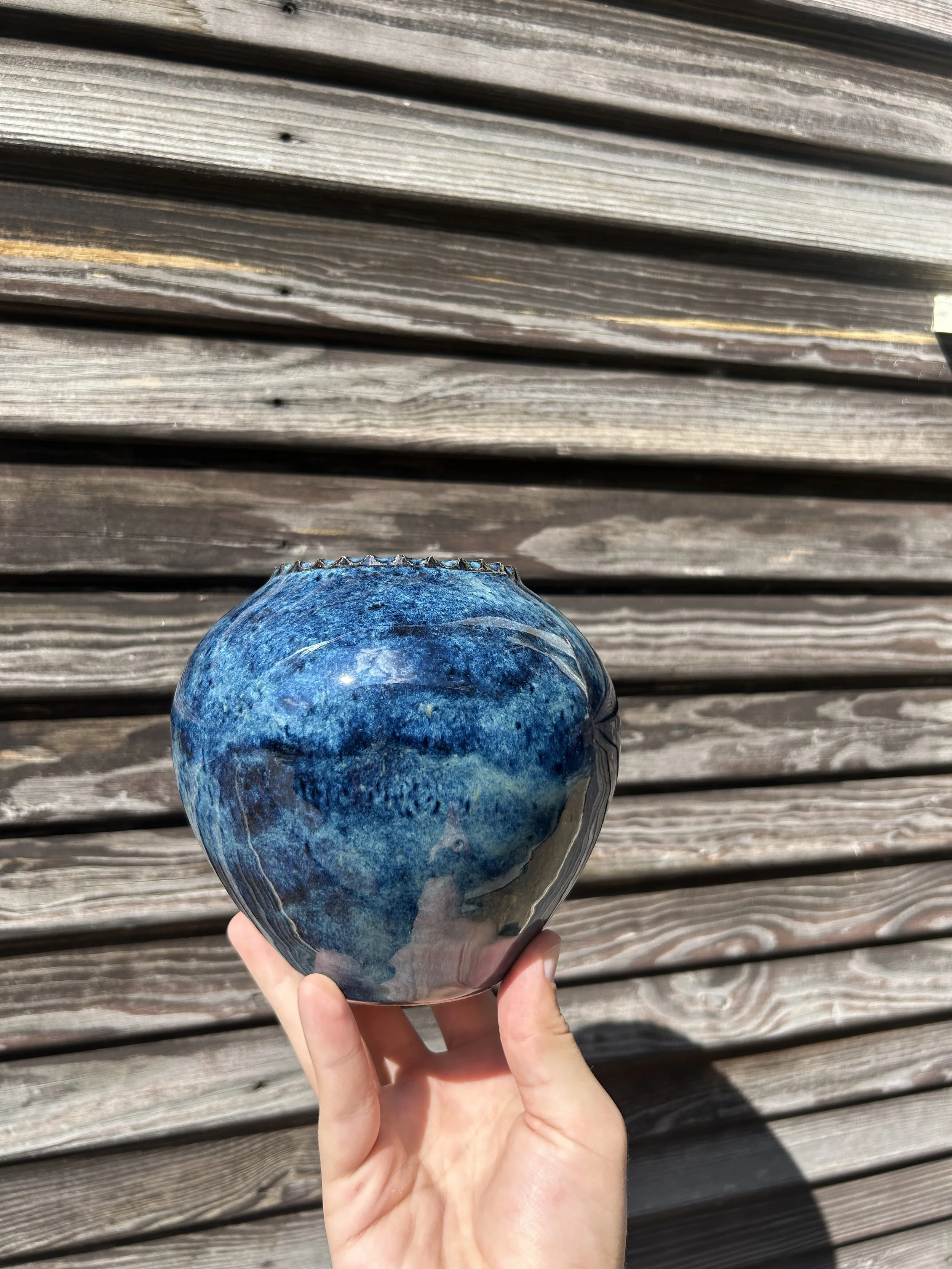 A person holding a vibrant blue and black patterned ceramic vase outdoors, with a weathered wooden surface in the background.