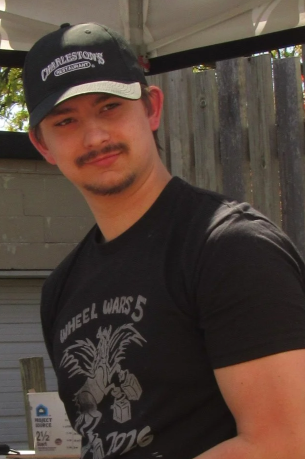 A young man with light skin, a mustache and goatee, wearing a black Cap with white text, black T-shirt with a graphic and text, outdoors in front of a wooden fence, under a white canopy.