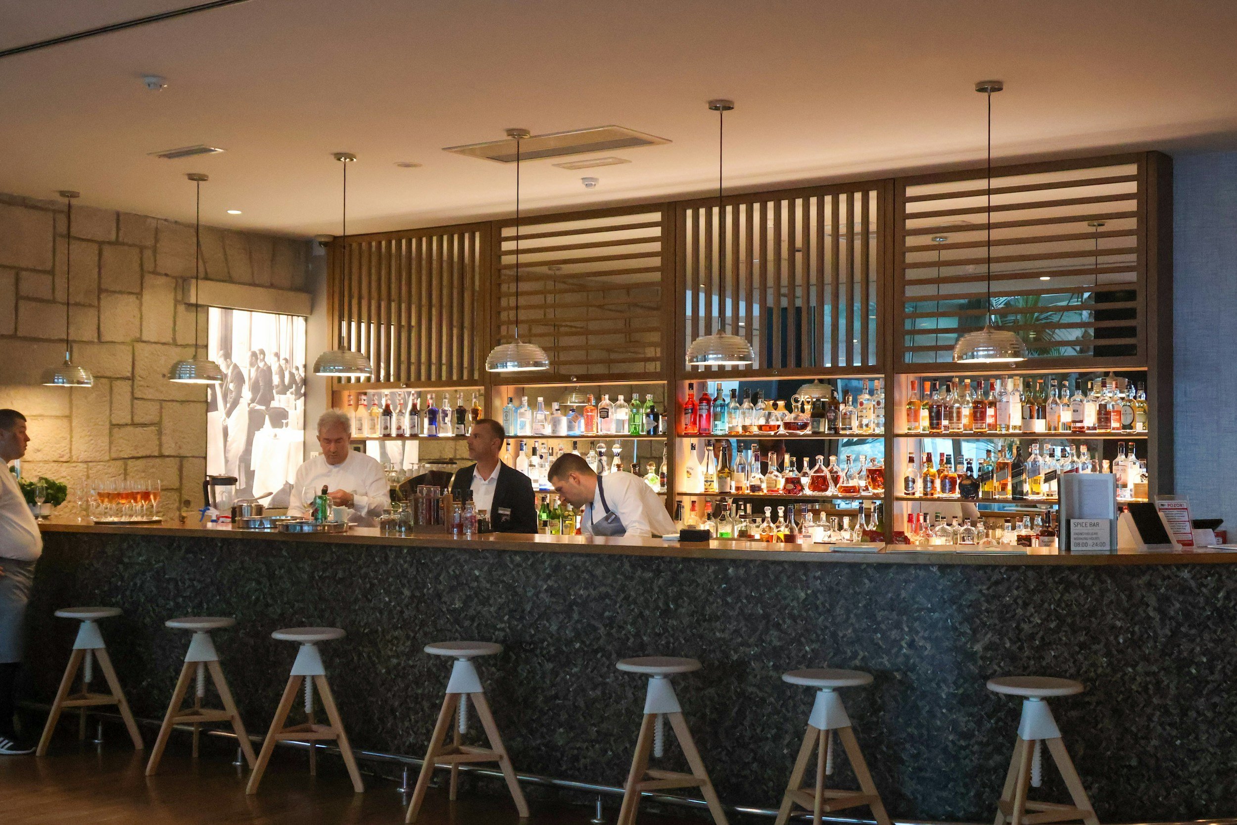 A bar area inside a restaurant with a stone wall, bar counter, and barstools. There are three men behind the bar preparing drinks, with a large assortment of liquor bottles on shelves behind them. Pendant lights hang from the ceiling, and there is a promotional sign on the right side of the counter.