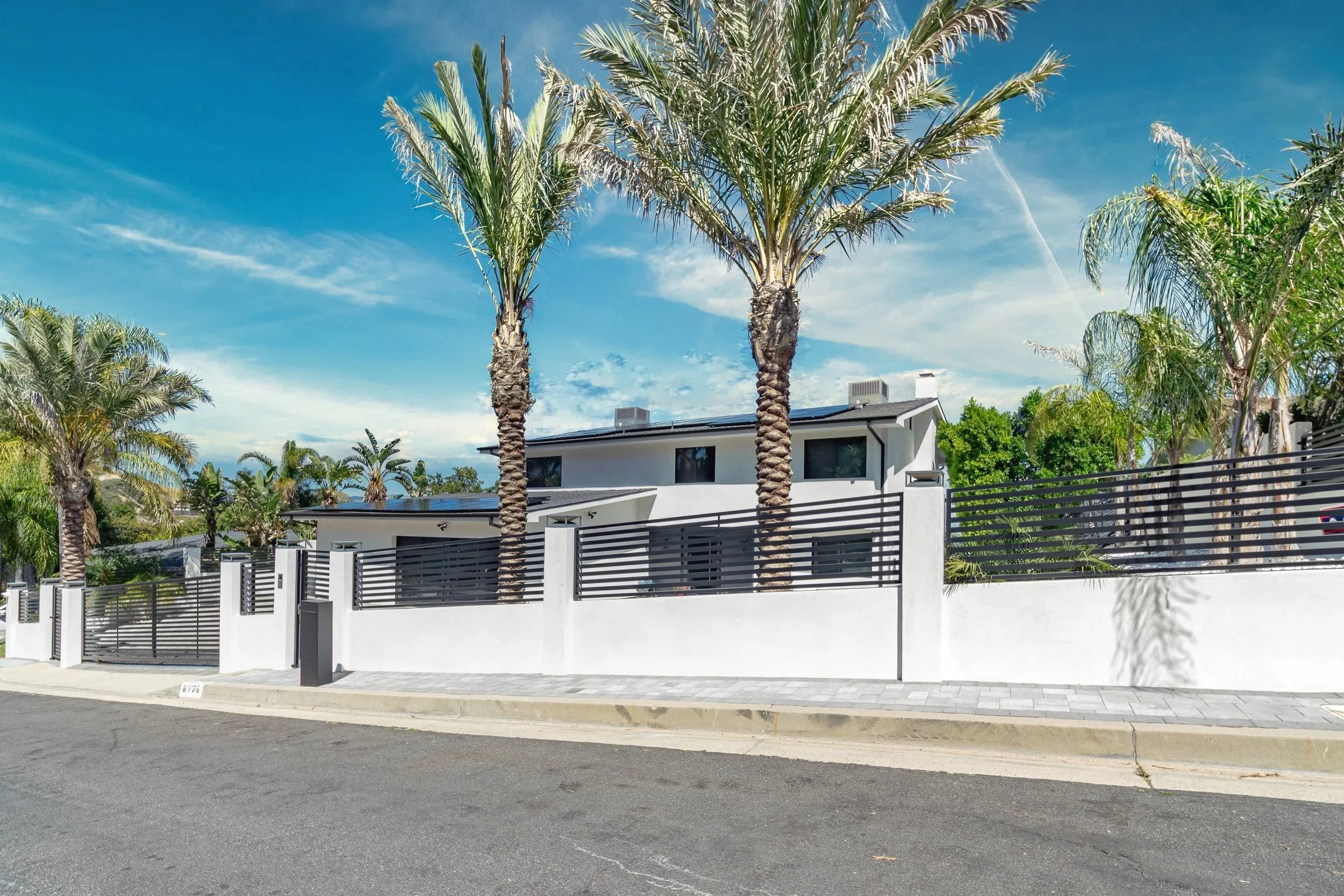 Modern white house with black fencing, palm trees, and a bright blue sky in the background.