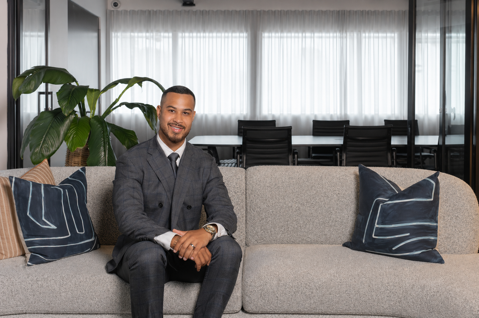 A well-dressed man in a gray suit and tie sitting on a beige sofa in a modern office lounge, with decorative pillows and a large green plant behind him, and a conference room with chairs and large windows with white curtains in the background.
