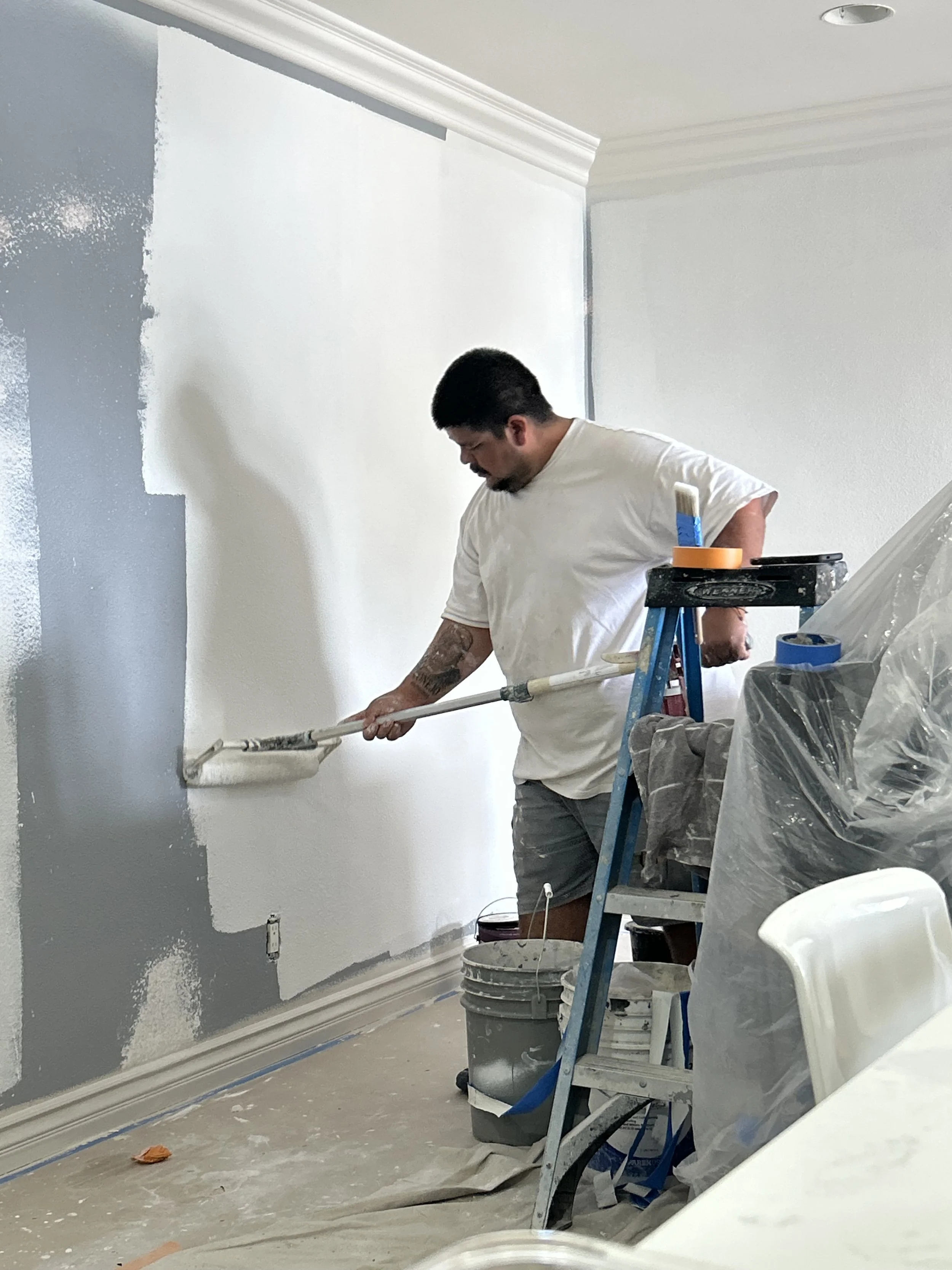 A man is painting a wall white using a roller while standing on a ladder. There are paint buckets, a painter's tarp, and painting supplies around him in a room under renovation.