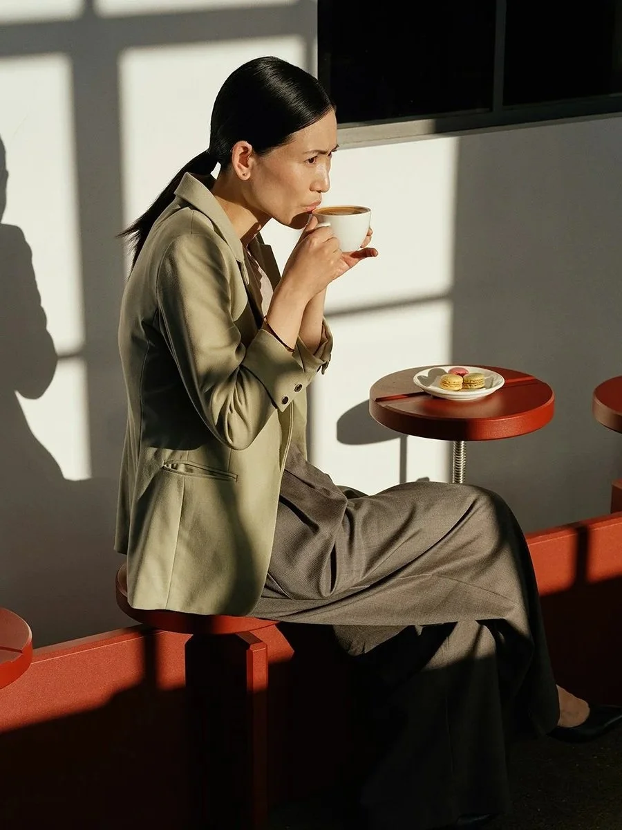 A woman with black hair tied back, wearing a beige blazer and gray pants, is sitting on a red bench, holding a cup of coffee or tea, and enjoying the aroma. A small round red table with a white plate of macarons is beside her, and sunlight casts shadows through a window.