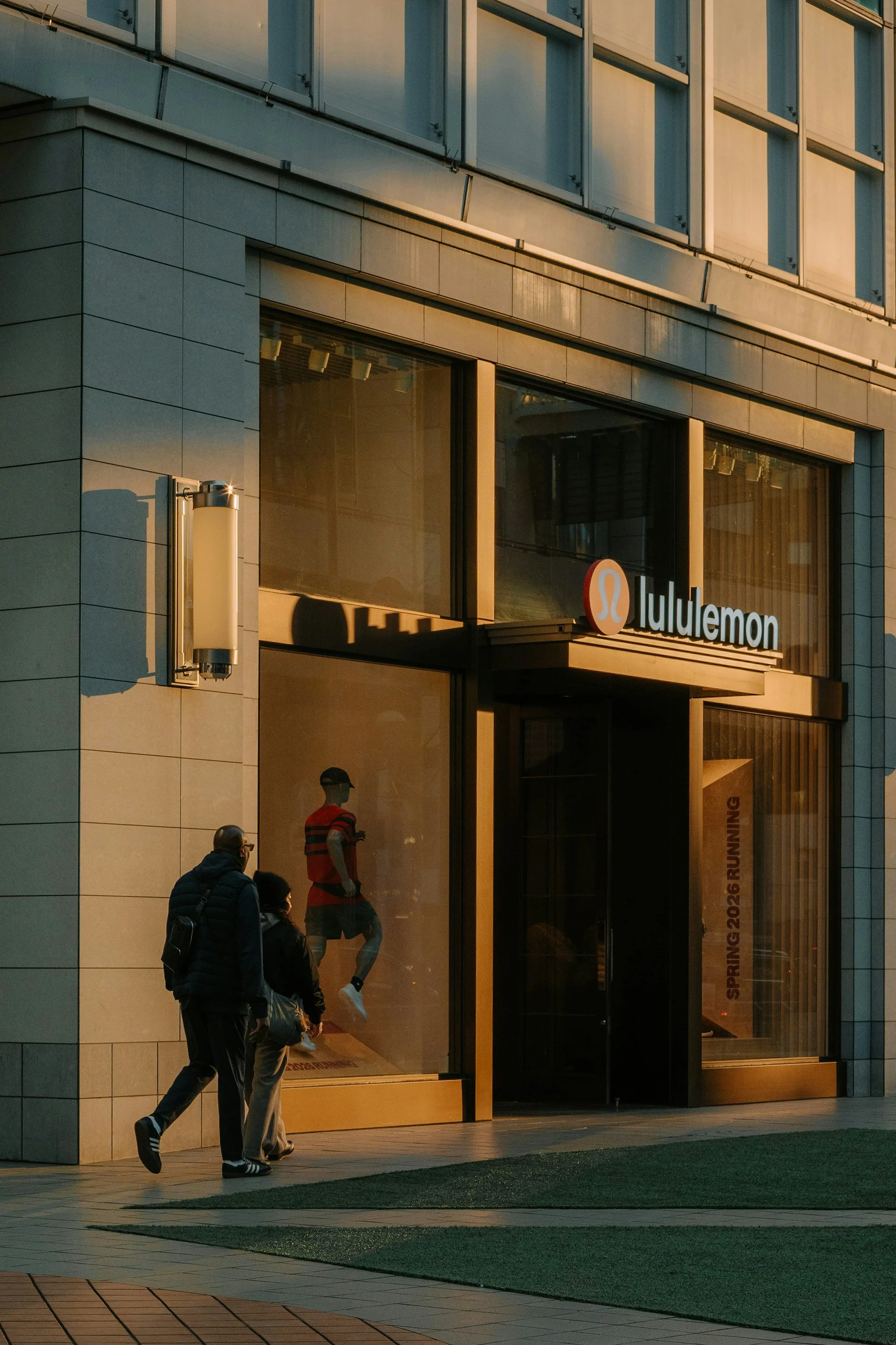 People walking past a retail store with a sign that reads 'lululemon' near the entrance, with large windows reflecting the surroundings and a gray tiled exterior.