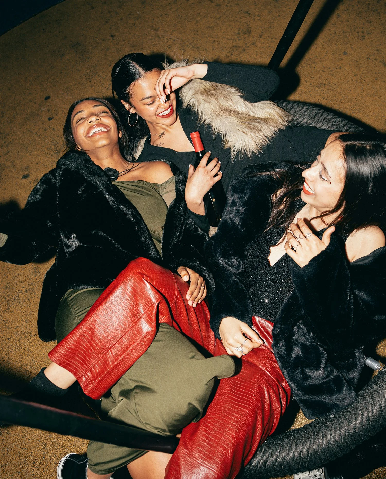 Three women are lying on the floor, smiling and laughing together. One woman is holding a bottle of wine. They are dressed stylishly, with one wearing red leather pants, another in black, and the third in olive green. They appear to be having fun at a social gathering.