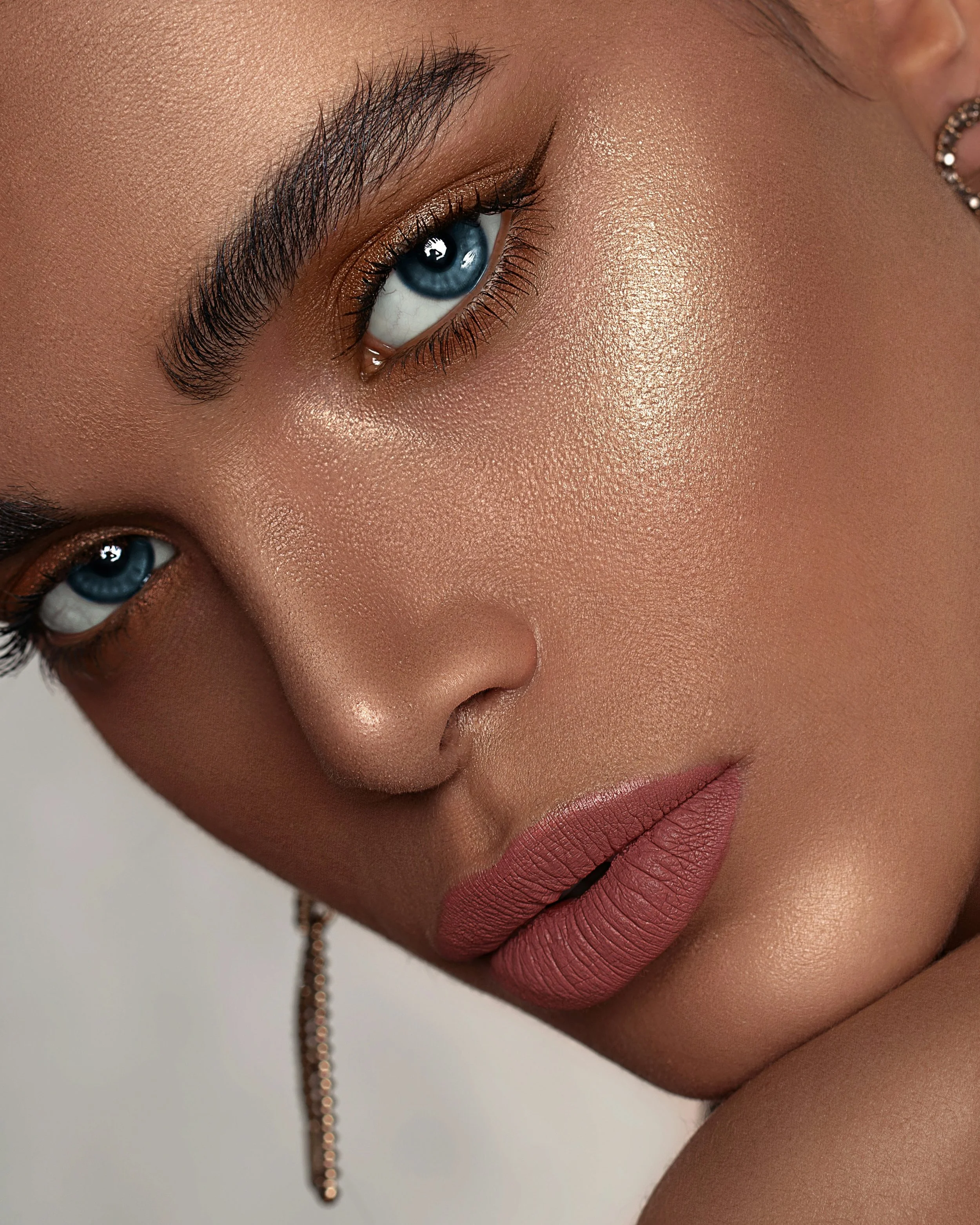 Close-up of a woman’s face showcasing detailed makeup, blue eye, textured skin, and natural lips, with visible earrings and a chain.
