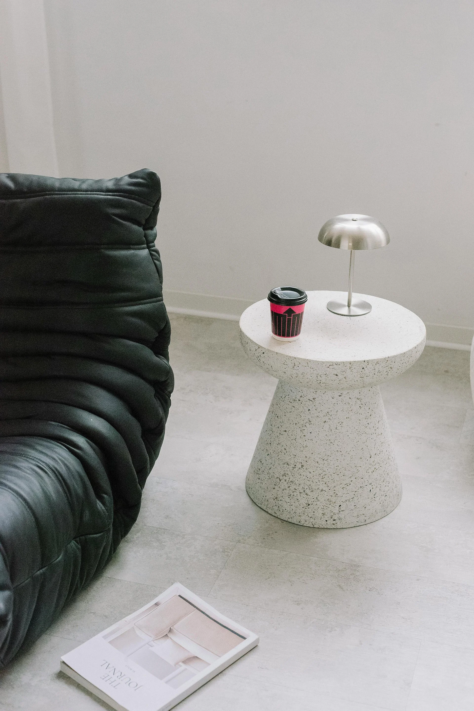 A black leather sofa with a tufted design next to a round speckled white side table. The table holds a metallic silver desk lamp and a black and pink travel mug. A magazine with a white cover featuring a minimalist interior scene is on the light-colored floor, with some sheets of paper nearby.