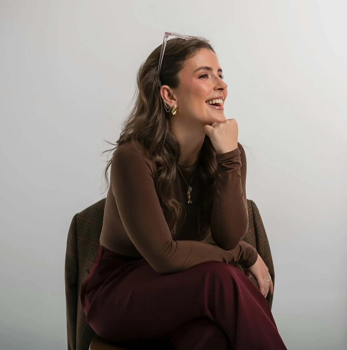 A woman with long wavy brown hair, gold hoop earrings, glasses on her head, smiling and sitting with her chin resting on her hand against a plain white background.