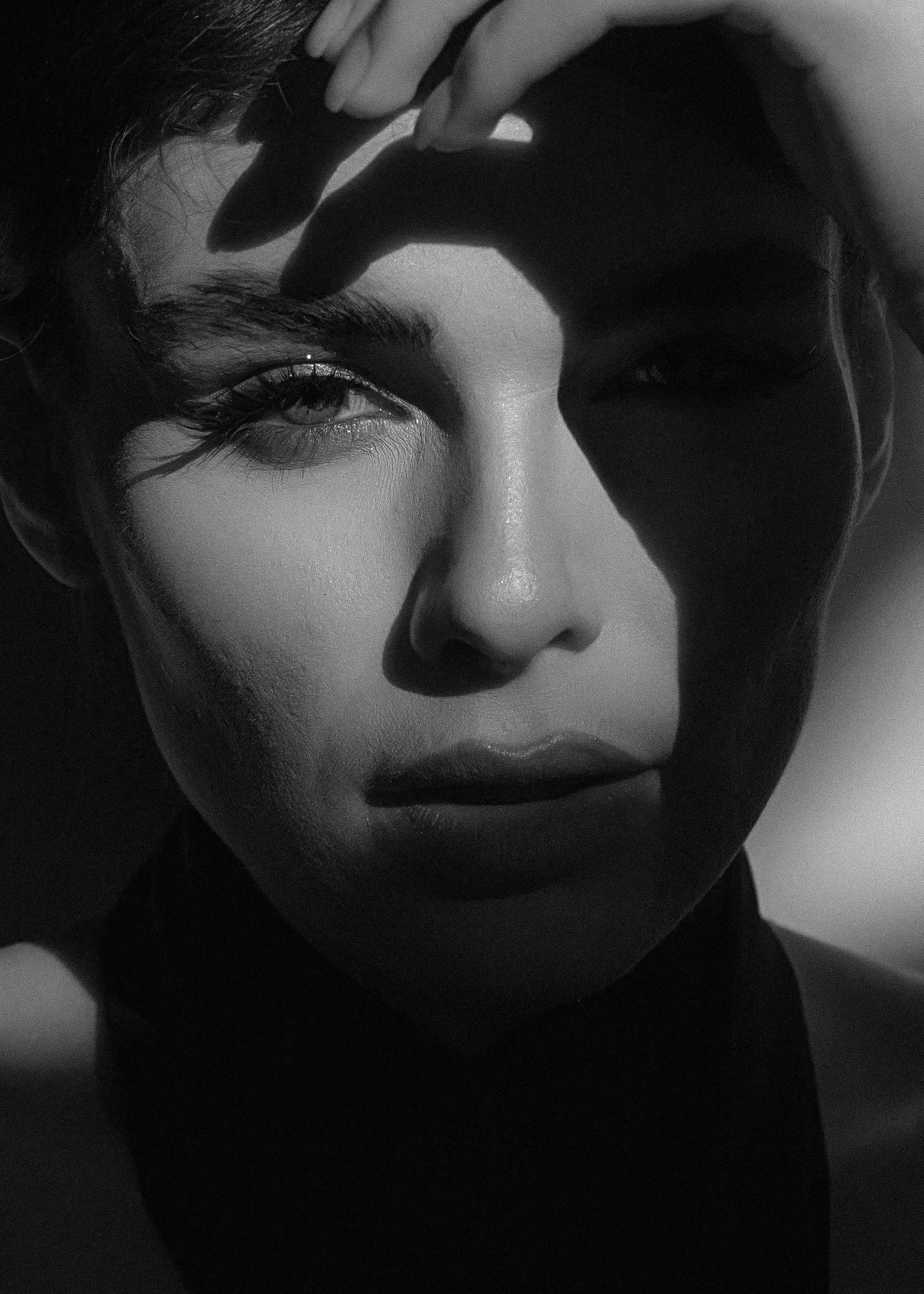 Black and white close-up of a person's face with one eye visible, hand touching forehead, dark hair, and makeup on lips.