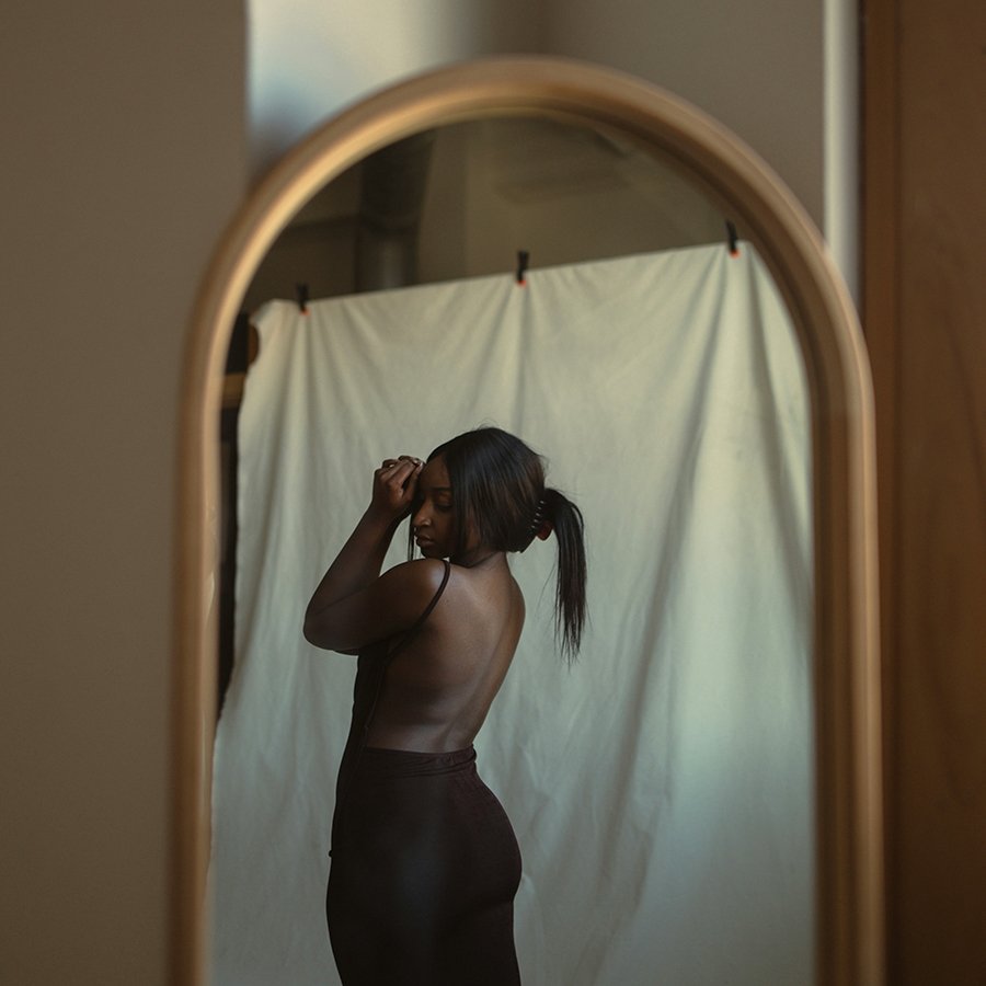 A woman with dark hair tied back in a ponytail taking a mirror selfie, showing her back and side profile, with a light-colored backdrop behind her.