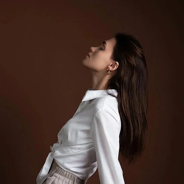 Side profile of a woman with long brown hair, wearing a white blouse and beige pants, against a brown background.