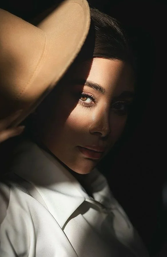 A woman with dark hair wearing a wide-brimmed tan hat and a white shirt, partially shadowed with sunlight illuminating one eye and part of her face.